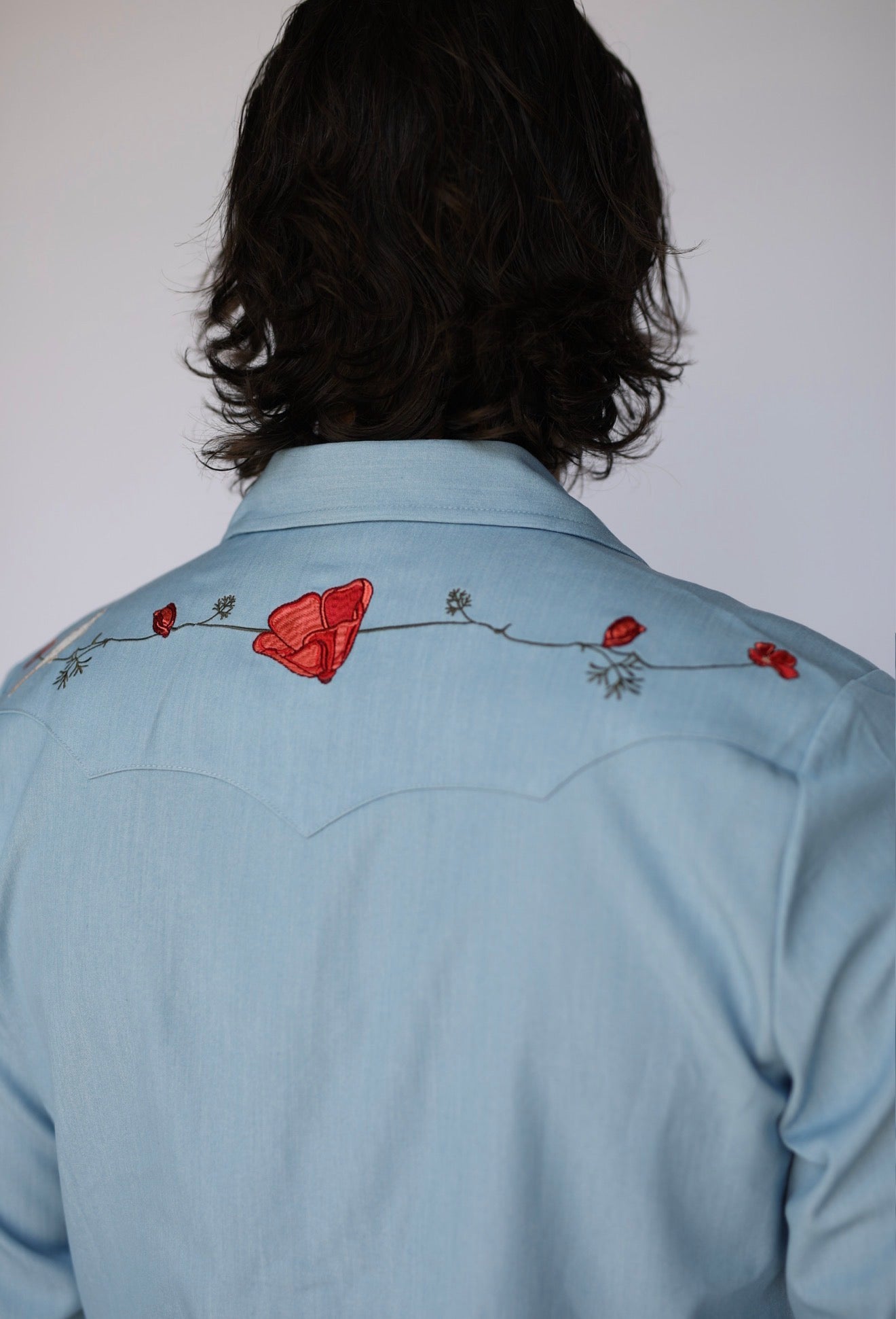 A person viewed from the back wearing a light blue western-style shirt with red embroidered poppy flowers and a floral vine across the upper back.