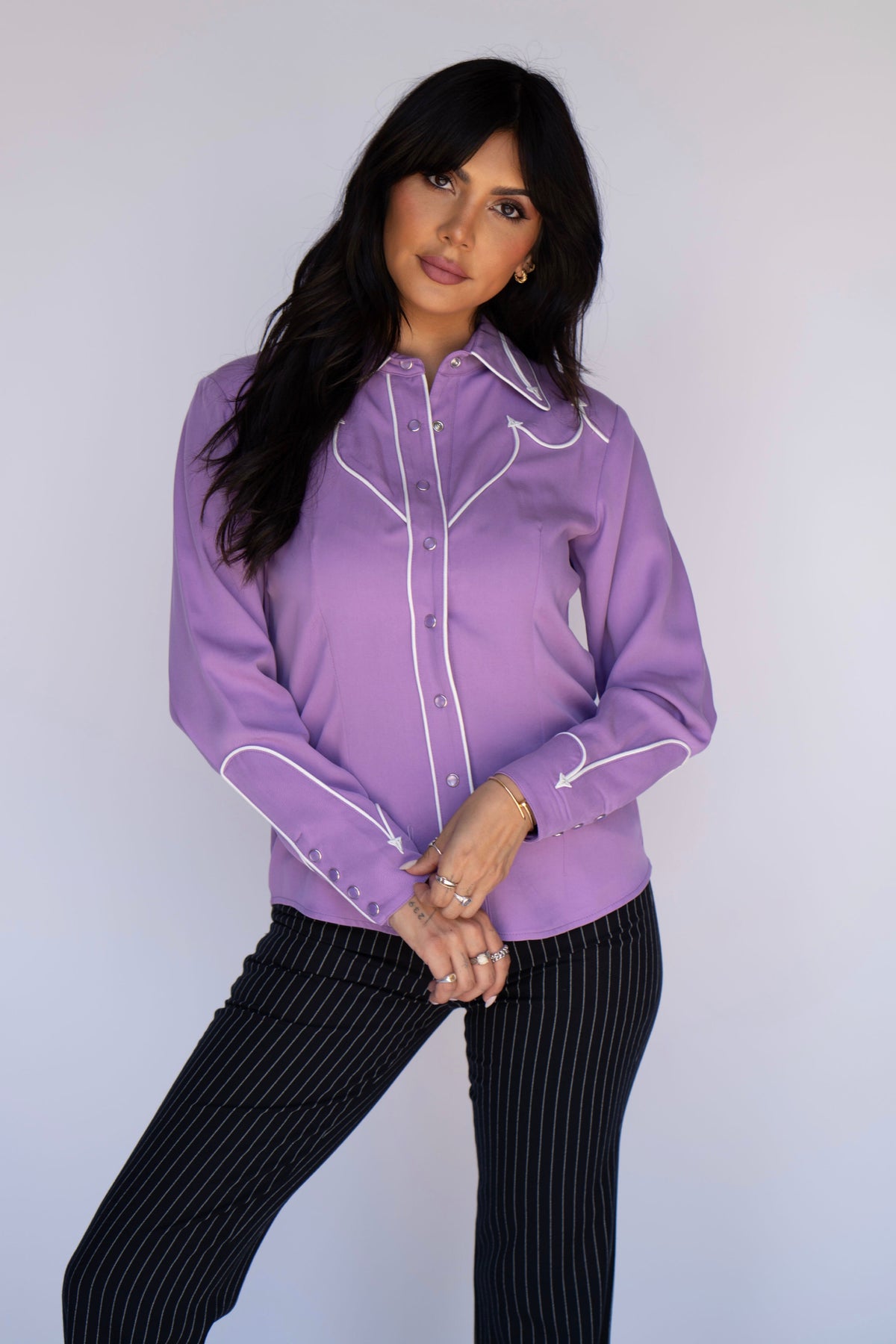 A woman modeling a lavender purple western-style long-sleeve button-up shirt with white contrast piping, paired with black pinstriped pants, against a plain light gray studio background.