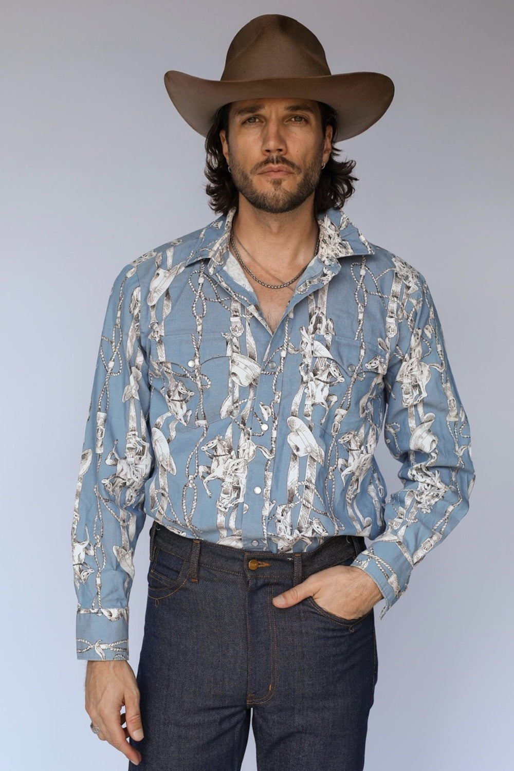 A man wearing a light blue long-sleeve western shirt with a white and gray cow print, a brown cowboy hat, and dark blue jeans, standing against a light gray studio background.