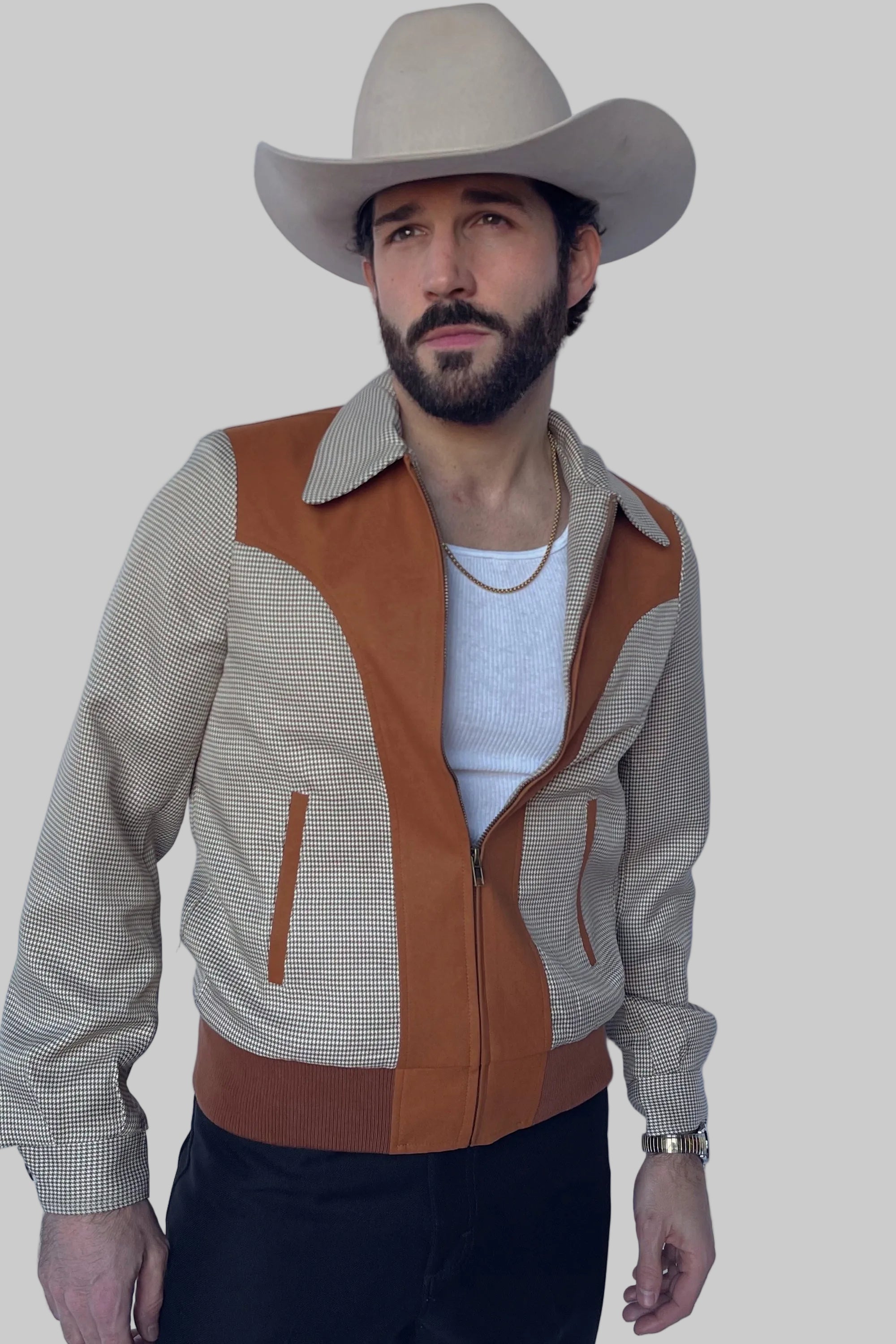 A man wearing a beige cowboy hat, a tan and beige striped zip-up jacket with brown suede-like panels, a white t-shirt, and black pants, standing against a light blue background.