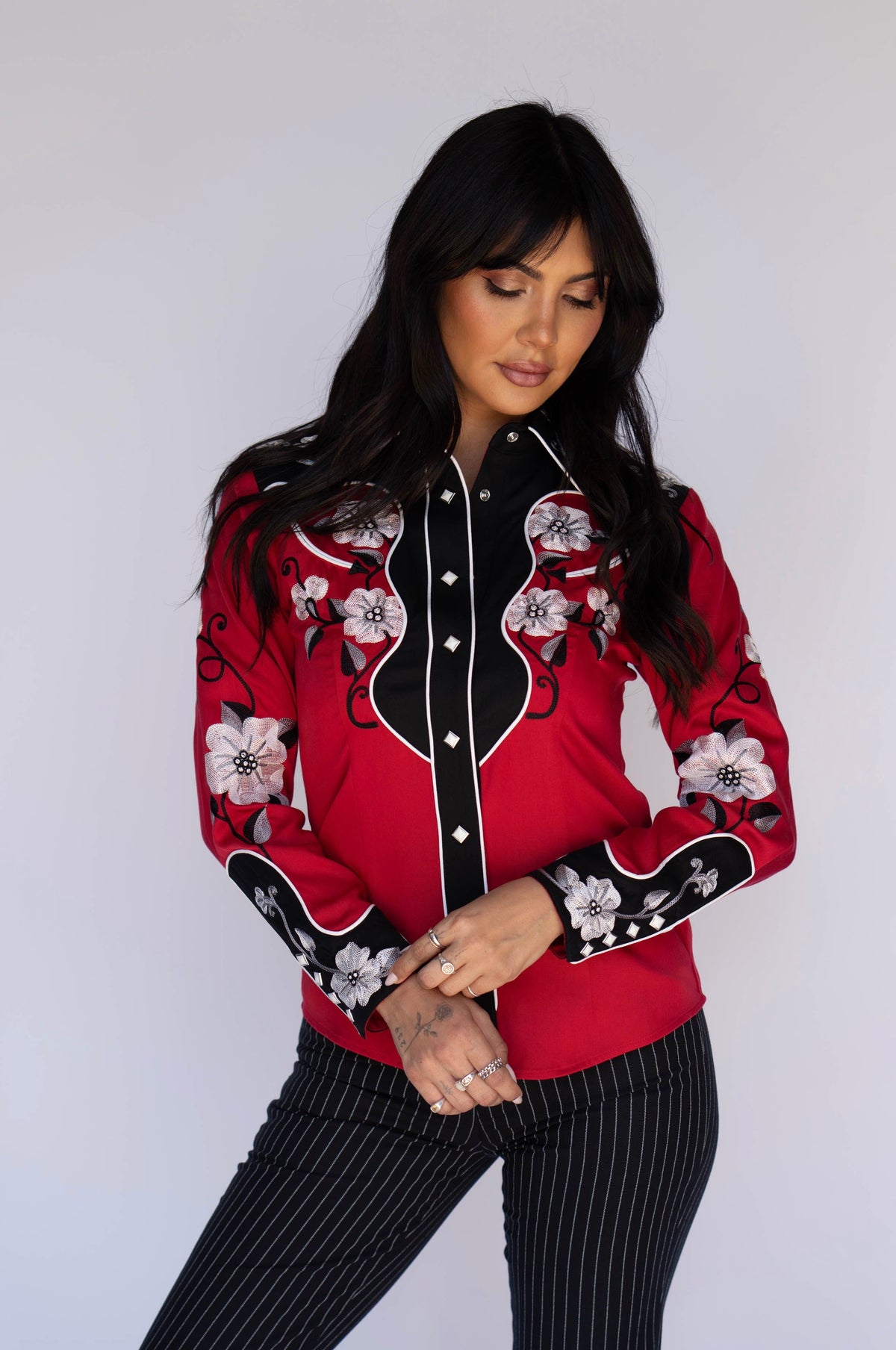 A woman modeling a red western-style button-up shirt with black panels, white floral embroidery, and black pinstriped pants against a plain light gray background.