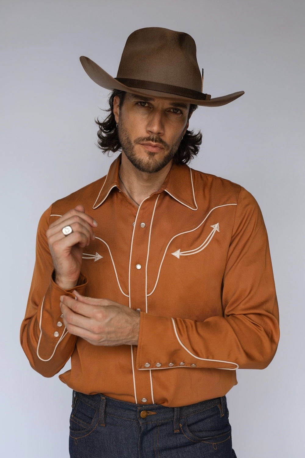 A man wearing a brown cowboy hat and a rust-orange western long-sleeve shirt with white contrast stitching, paired with dark blue jeans, against a light gray studio background.