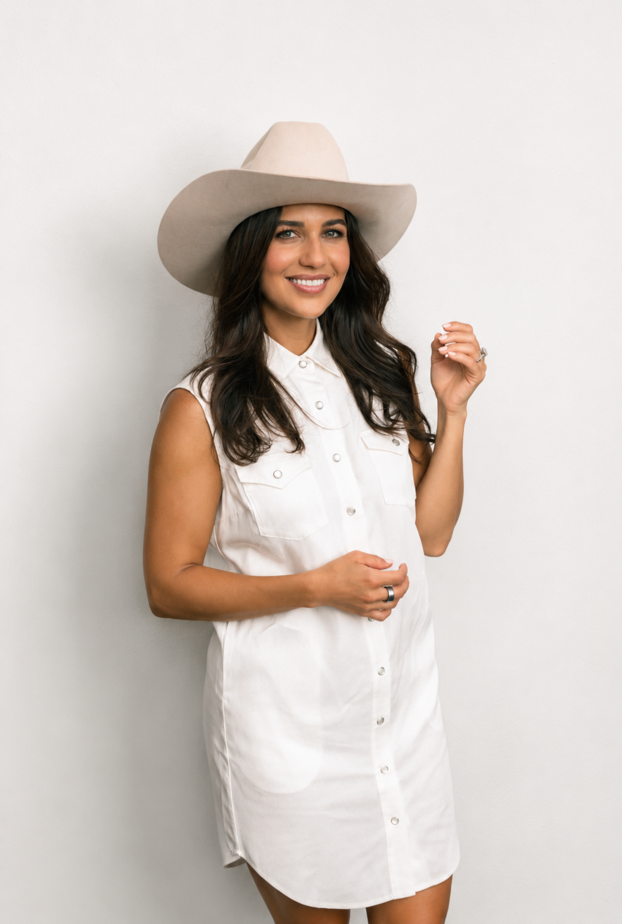A woman wearing a white sleeveless button-front shirt dress and a light beige cowboy hat, standing against a plain light gray studio background.