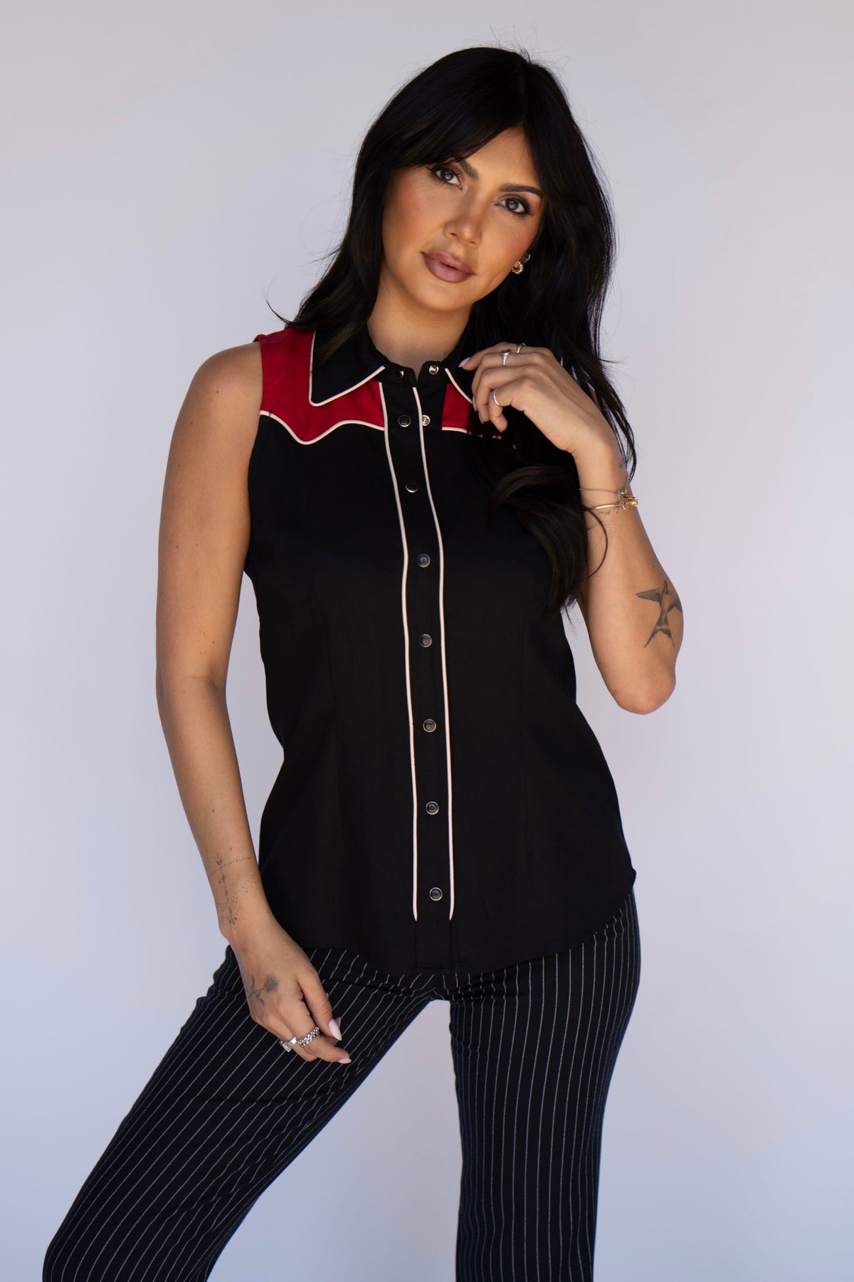 A woman modeling a sleeveless black button-front top with red and white western-style collar and trim details, paired with black pinstriped pants, against a plain light gray studio background.