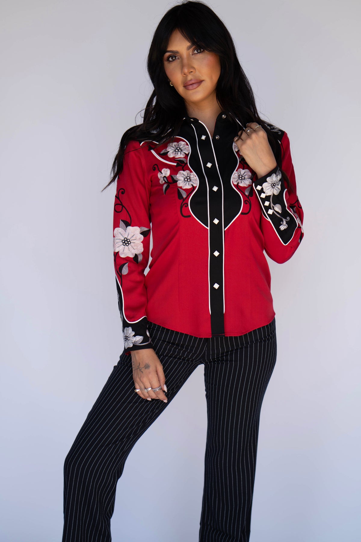 A woman modeling a red western-style button-up shirt with black panels, white piping, and floral embroidery on the sleeves, paired with black pinstriped pants against a light gray studio background.