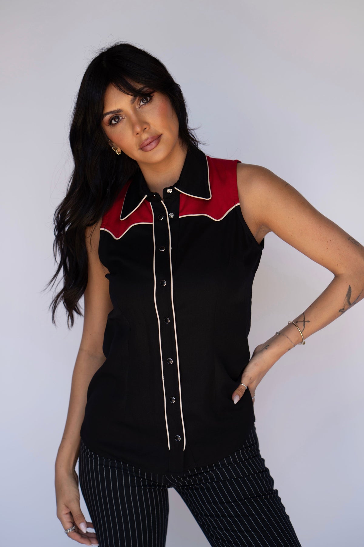 A woman modeling a sleeveless black button-front top with a red yoke and cream piping details, paired with black and white pinstriped pants, against a plain light gray background.