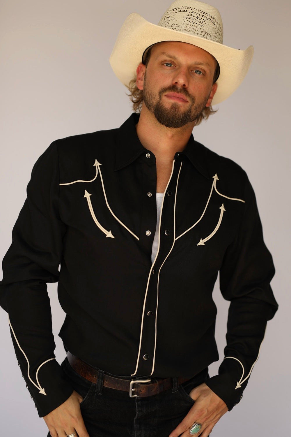 A man wearing a black western-style button-up shirt with white embroidered arrow details on the chest and sleeves, paired with a cream cowboy hat, dark jeans, and a brown belt, posed against a light gray studio background.