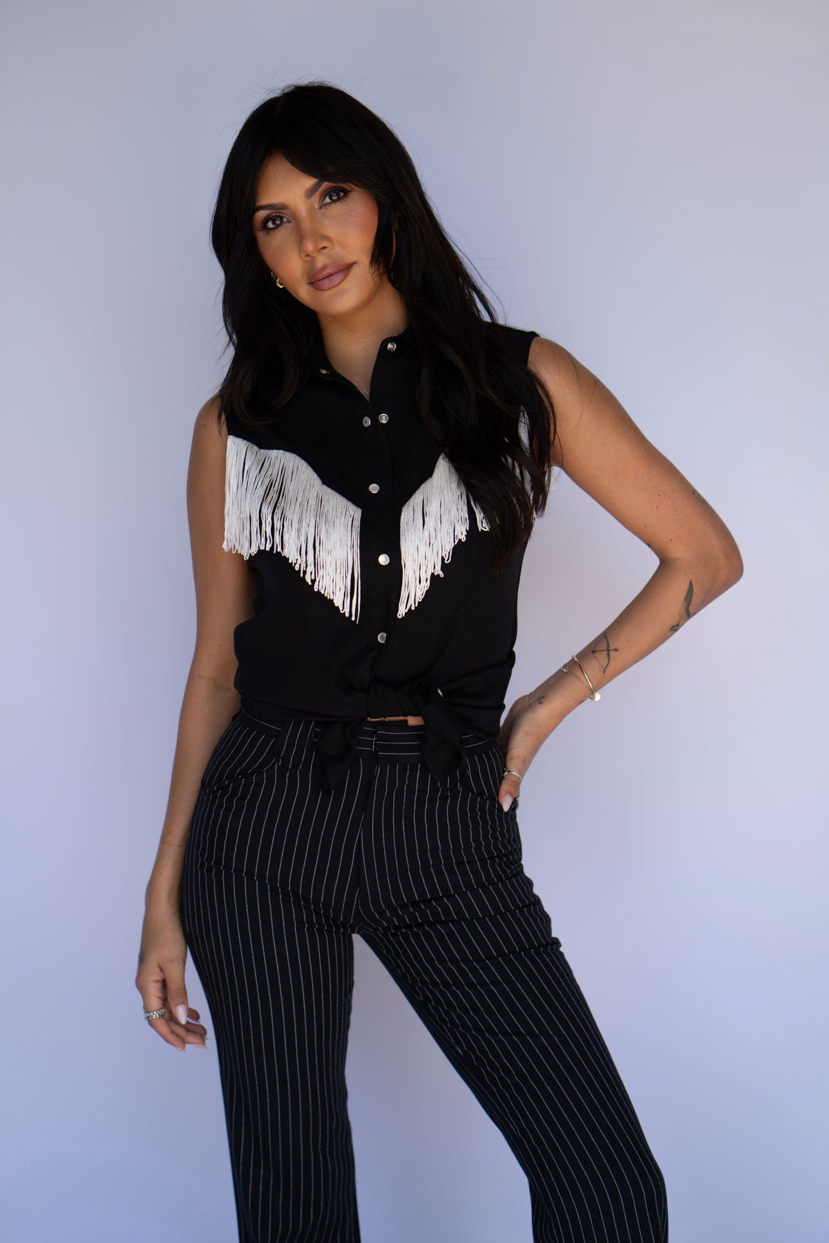 A woman modeling a black sleeveless button-up shirt with white fringe across the chest, paired with black pinstriped trousers, against a light gray studio background.