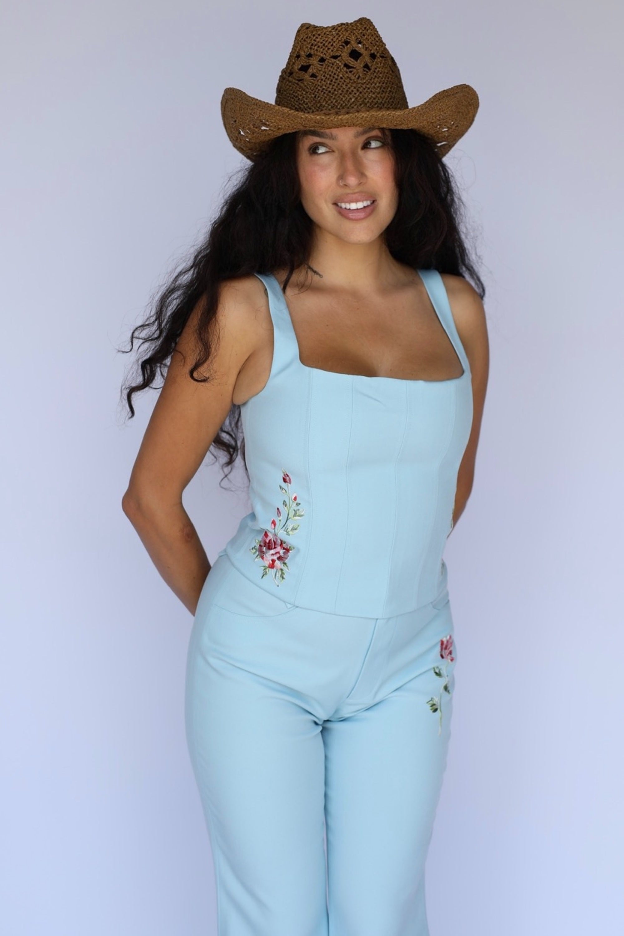 A woman modeling a light blue sleeveless jumpsuit with floral embroidery on the sides, paired with a brown woven cowboy hat, against a plain light gray studio background.
