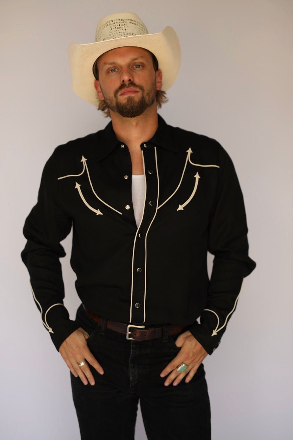 A man wearing a black western-style button-up shirt with cream embroidery, a white cowboy hat, and dark jeans, standing against a plain light background.