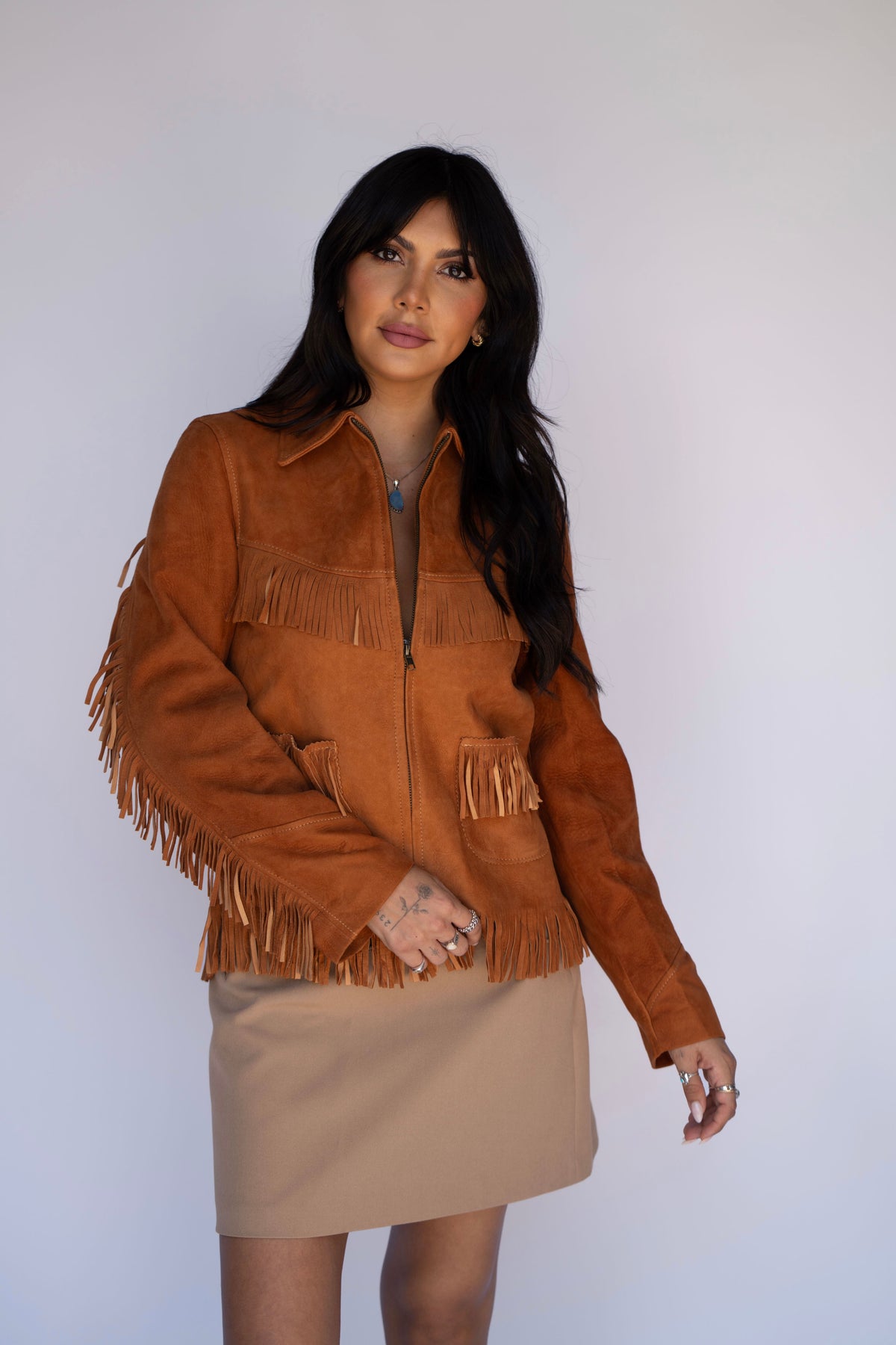 A woman modeling a brown suede fringe jacket with a half-zip front, paired with a beige mini skirt, against a plain light gray background.