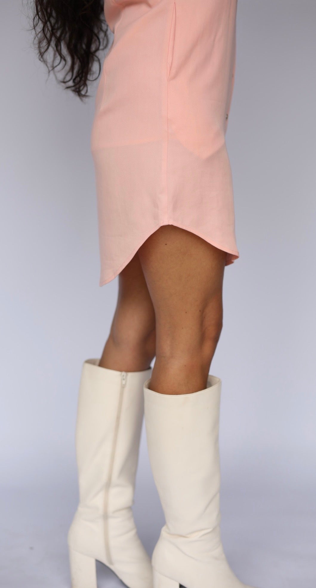 A person wearing a light pink skirt and off-white knee-high heeled boots, shown from the waist down against a plain light gray studio background.
