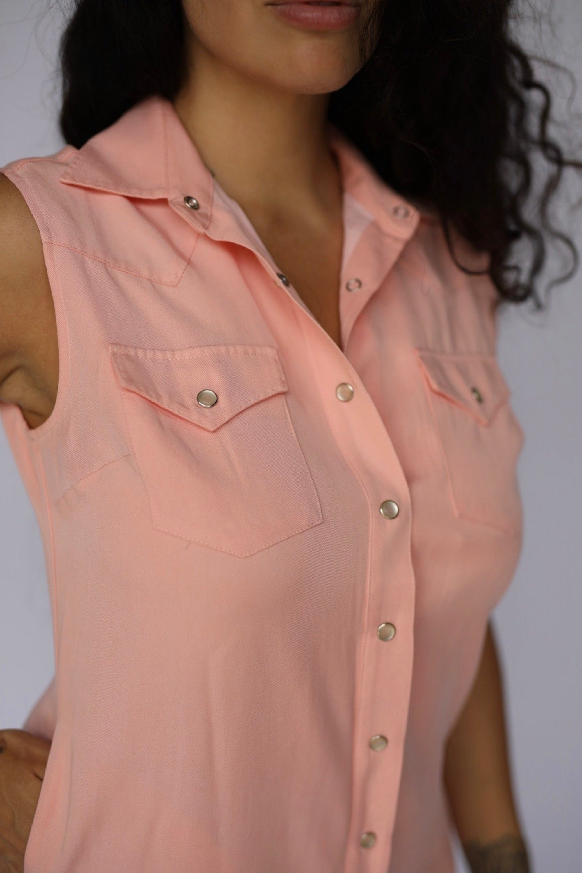 A close-up of a woman wearing a sleeveless, light pink button-down shirt with silver snap buttons and chest pockets.