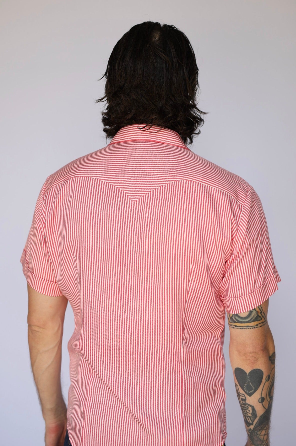 A man viewed from behind wearing a short-sleeve button-up shirt with thin red and white vertical stripes, set against a plain light gray background.
