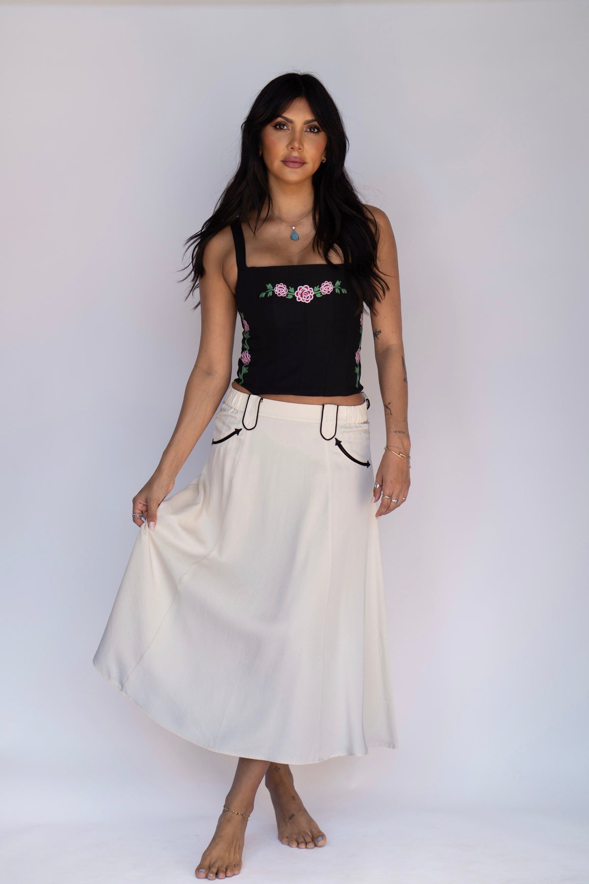 A woman modeling a black sleeveless crop top with pink and green floral embroidery and a high-waisted off-white maxi skirt with black drawstring details, standing against a plain white studio background.