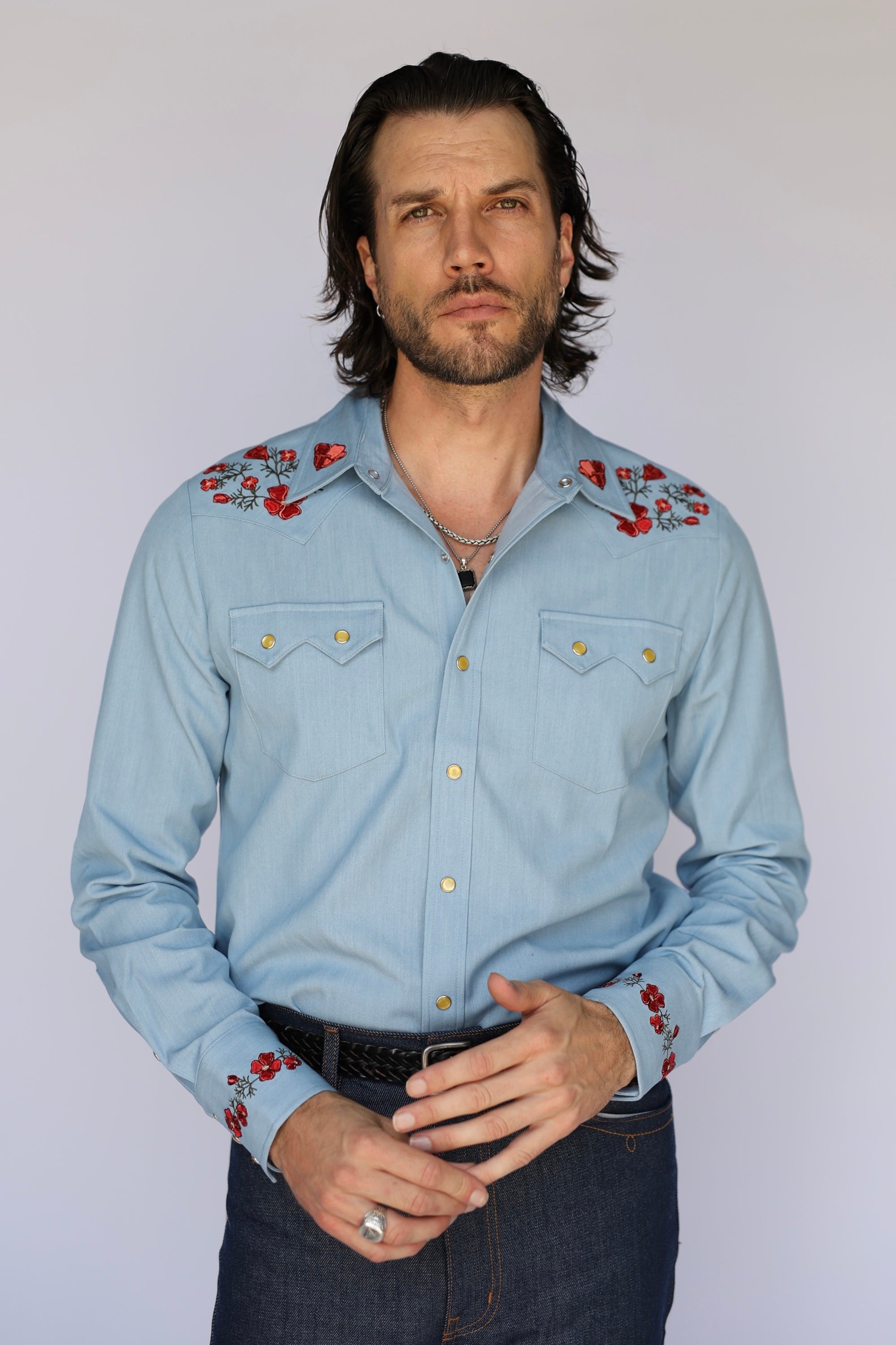 A man wearing a light blue western-style long-sleeve button-up shirt with red floral embroidery on the shoulders and cuffs, paired with dark jeans, against a plain light gray background.