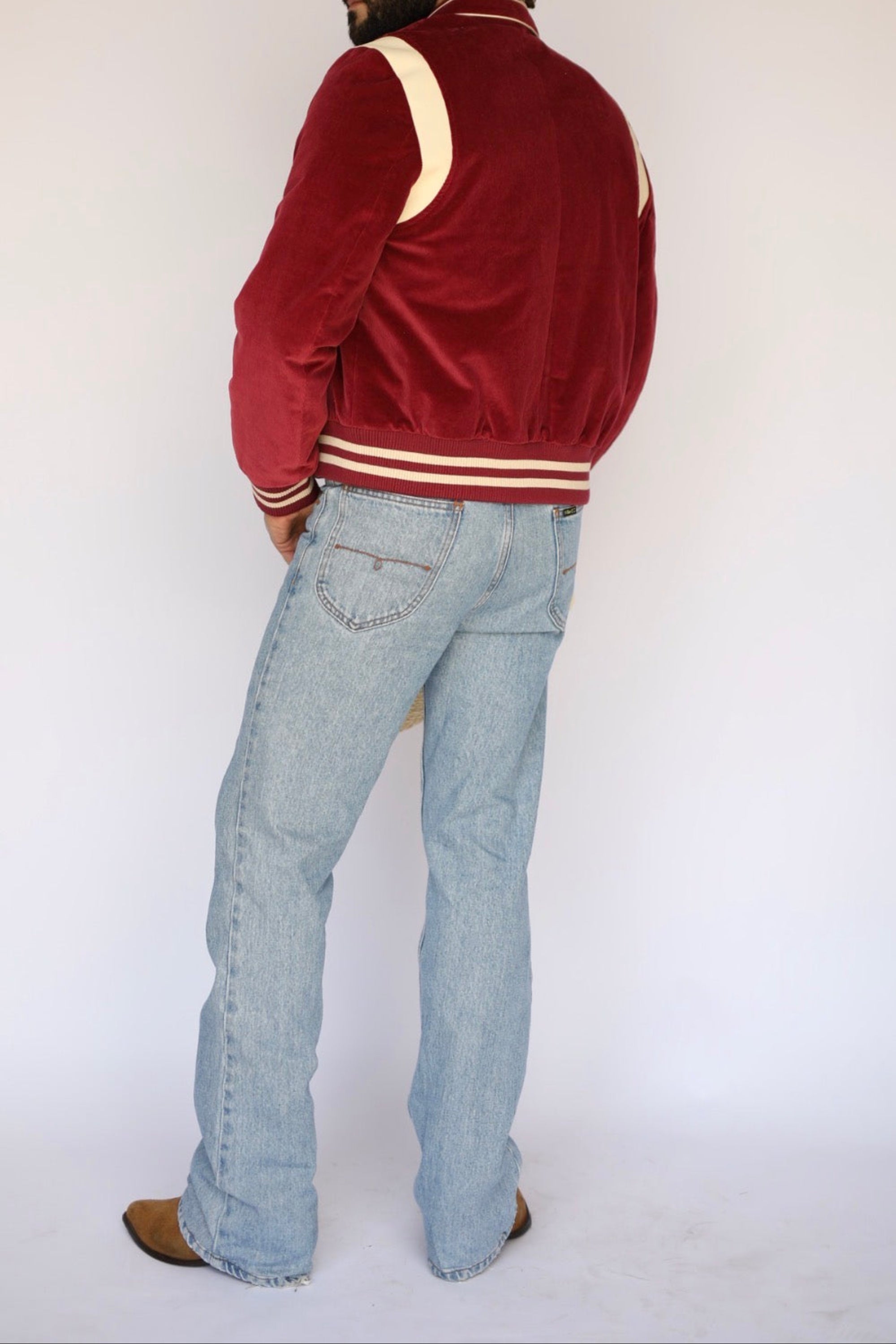 A man wearing a burgundy velvet varsity-style jacket with cream stripes, paired with light blue jeans and brown shoes, standing against a plain white studio background.