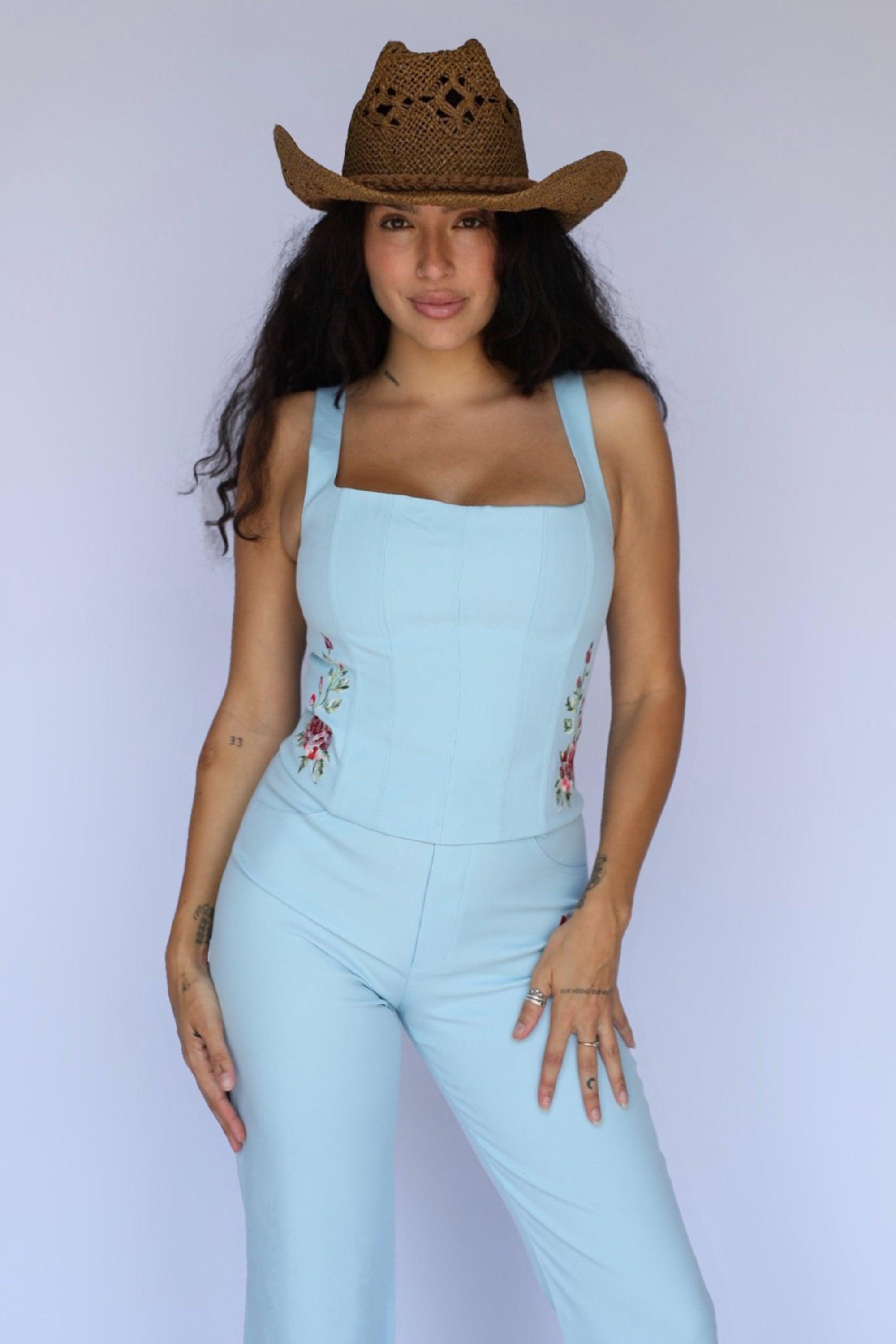 A woman modeling a light blue sleeveless jumpsuit with floral embroidery on the chest and sides, paired with a brown woven cowboy hat, against a plain light gray studio background.