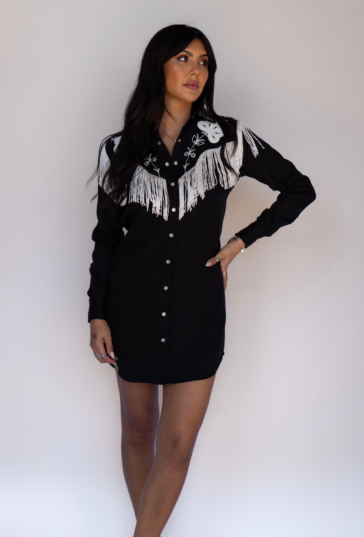 A woman modeling a black long-sleeve western-style mini shirt dress with white fringe and embroidery on the chest, photographed against a plain light background.