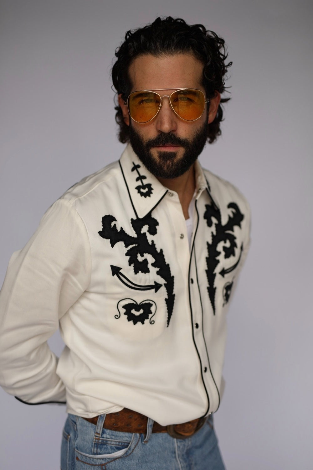 A man wearing a white western-style button-up shirt with bold black embroidered patterns, blue jeans, and gold-tinted aviator sunglasses, posed against a plain light gray studio background.