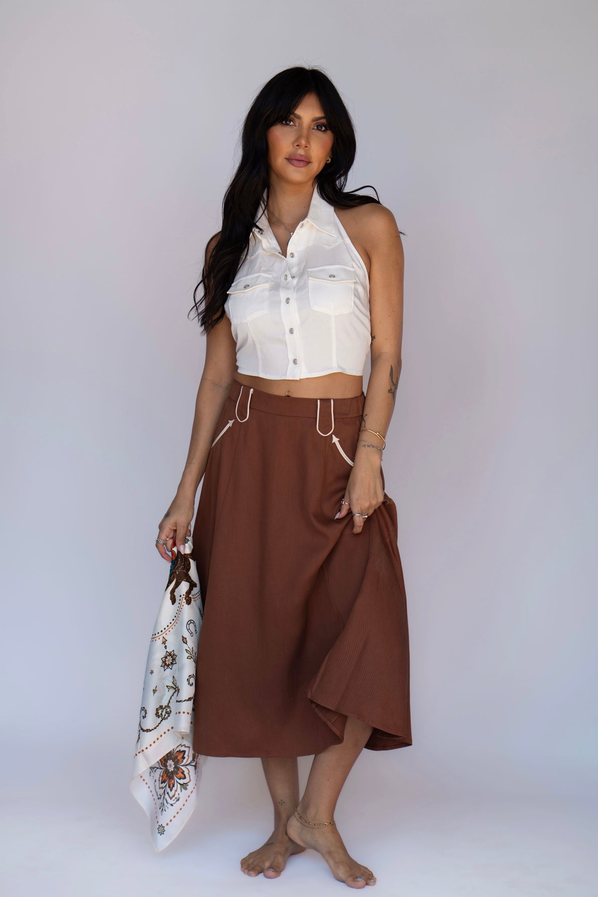 A woman stands barefoot against a plain light gray studio background, wearing a sleeveless white cropped button-front top and a brown midi skirt with white drawstrings. She holds a white patterned scarf.