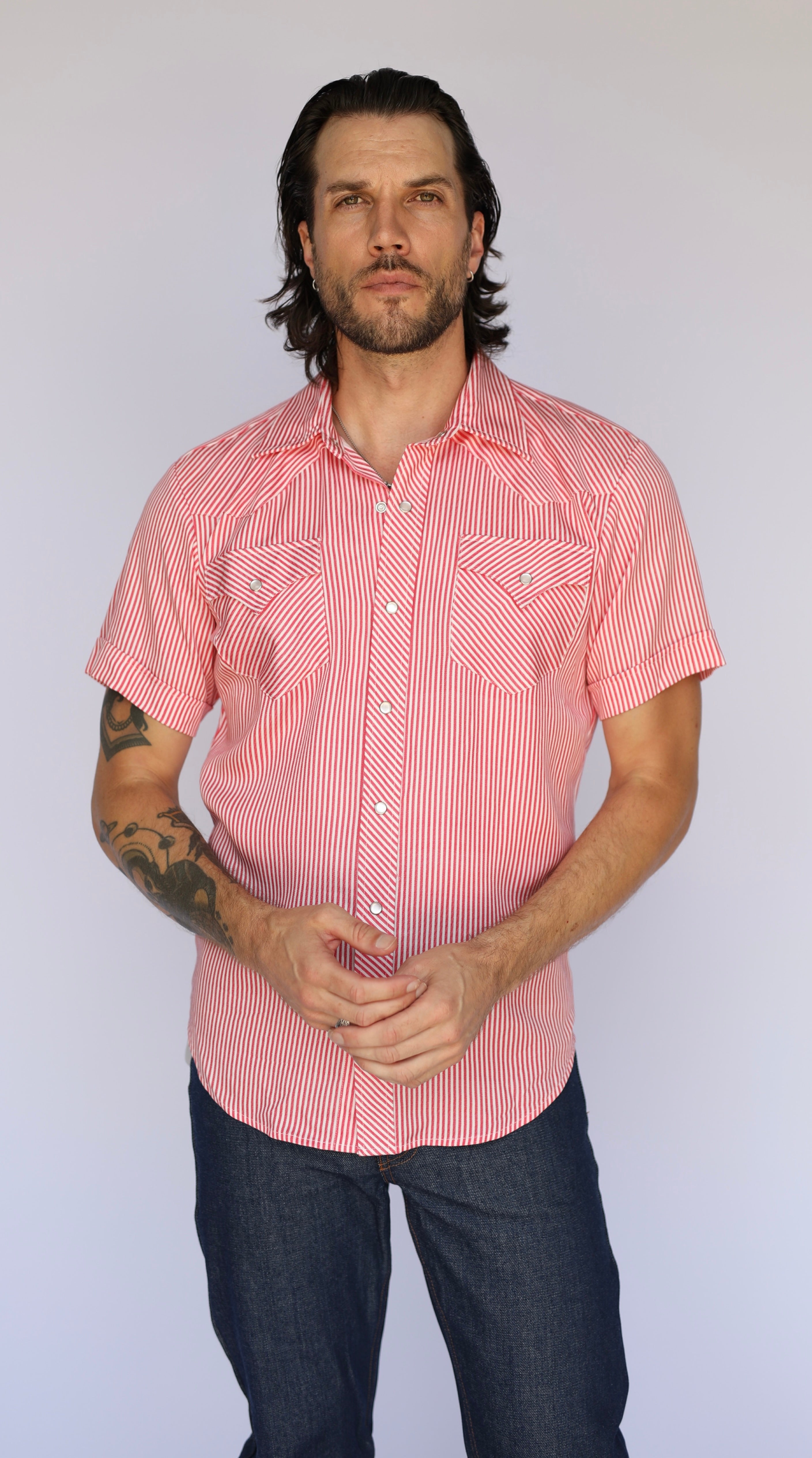 A man wearing a short-sleeve button-up shirt with a pink and white vertical stripe pattern, paired with dark jeans, standing against a plain light gray background.