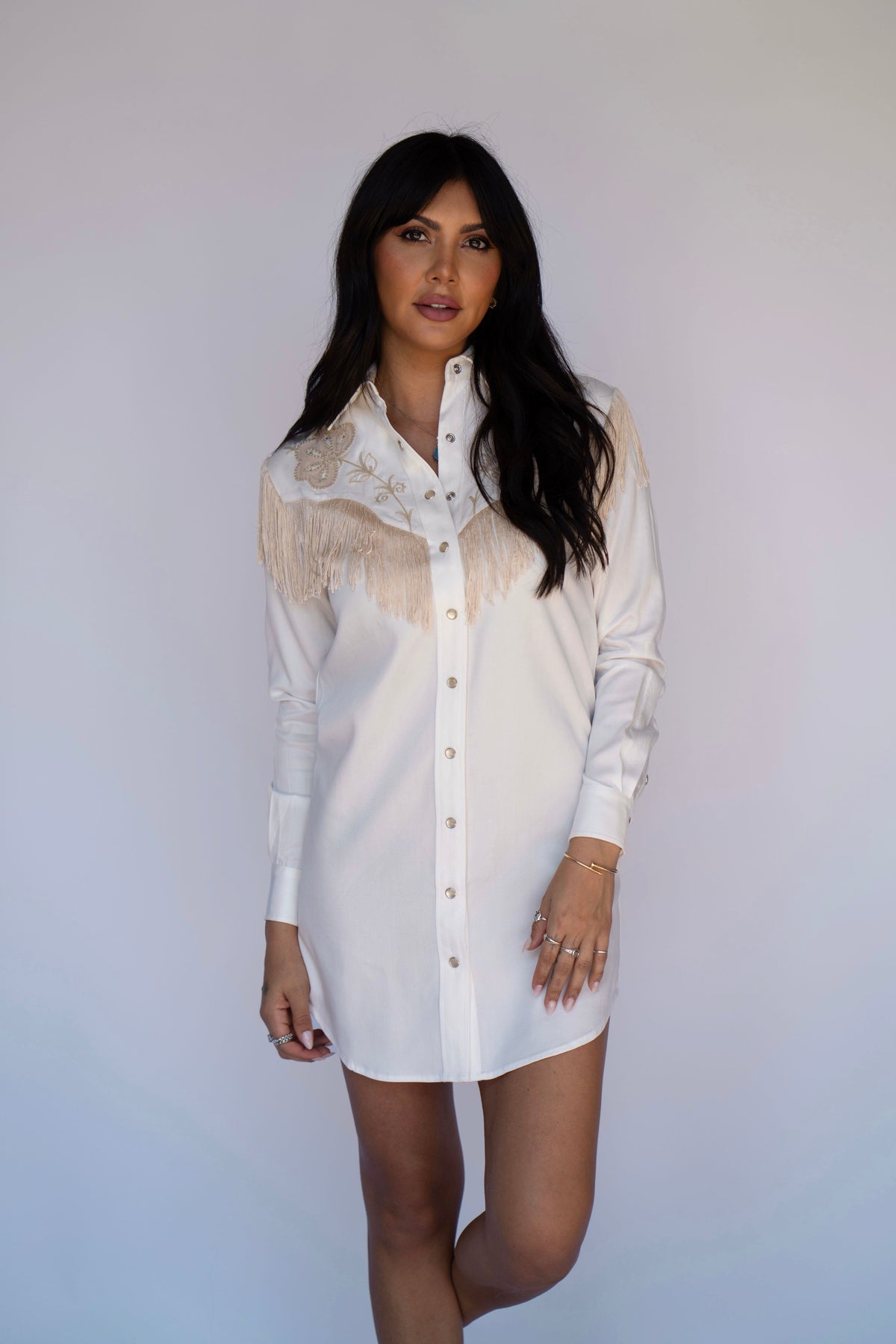 A woman modeling a white long-sleeve button-up shirt dress with beige fringe and embroidery on the chest, set against a plain light gray studio background.