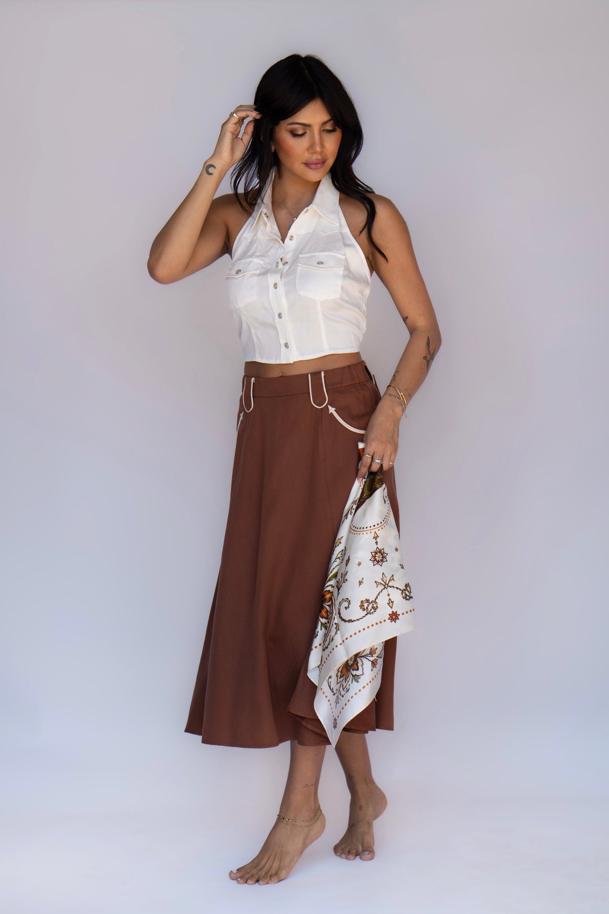 A woman stands barefoot against a plain light gray studio background, wearing a white sleeveless cropped top and a brown midi skirt, while holding a white patterned scarf.