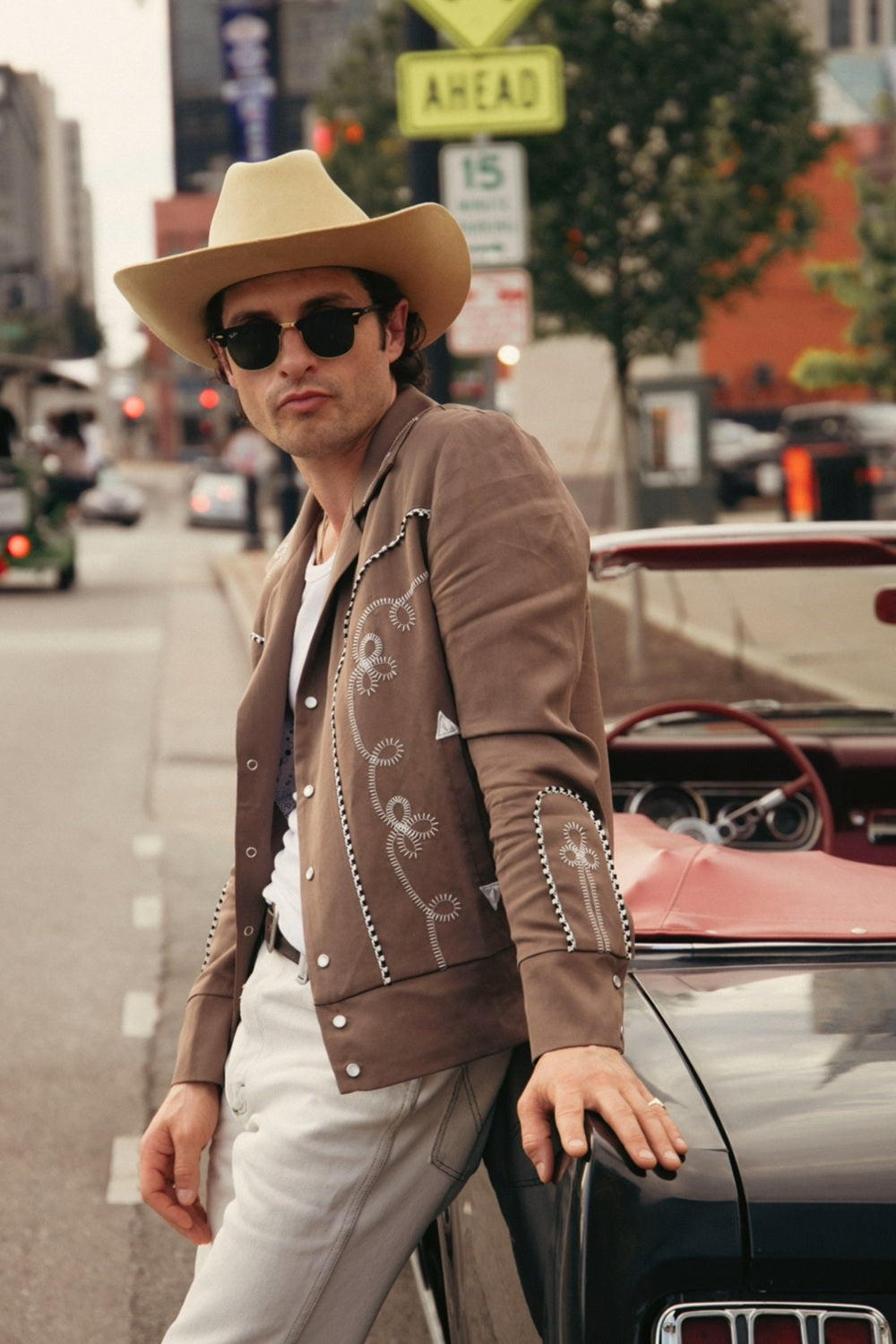 A man wearing a tan cowboy hat, brown western-style jacket with white embroidery, white shirt, and white pants leans against a vintage car on a city street.