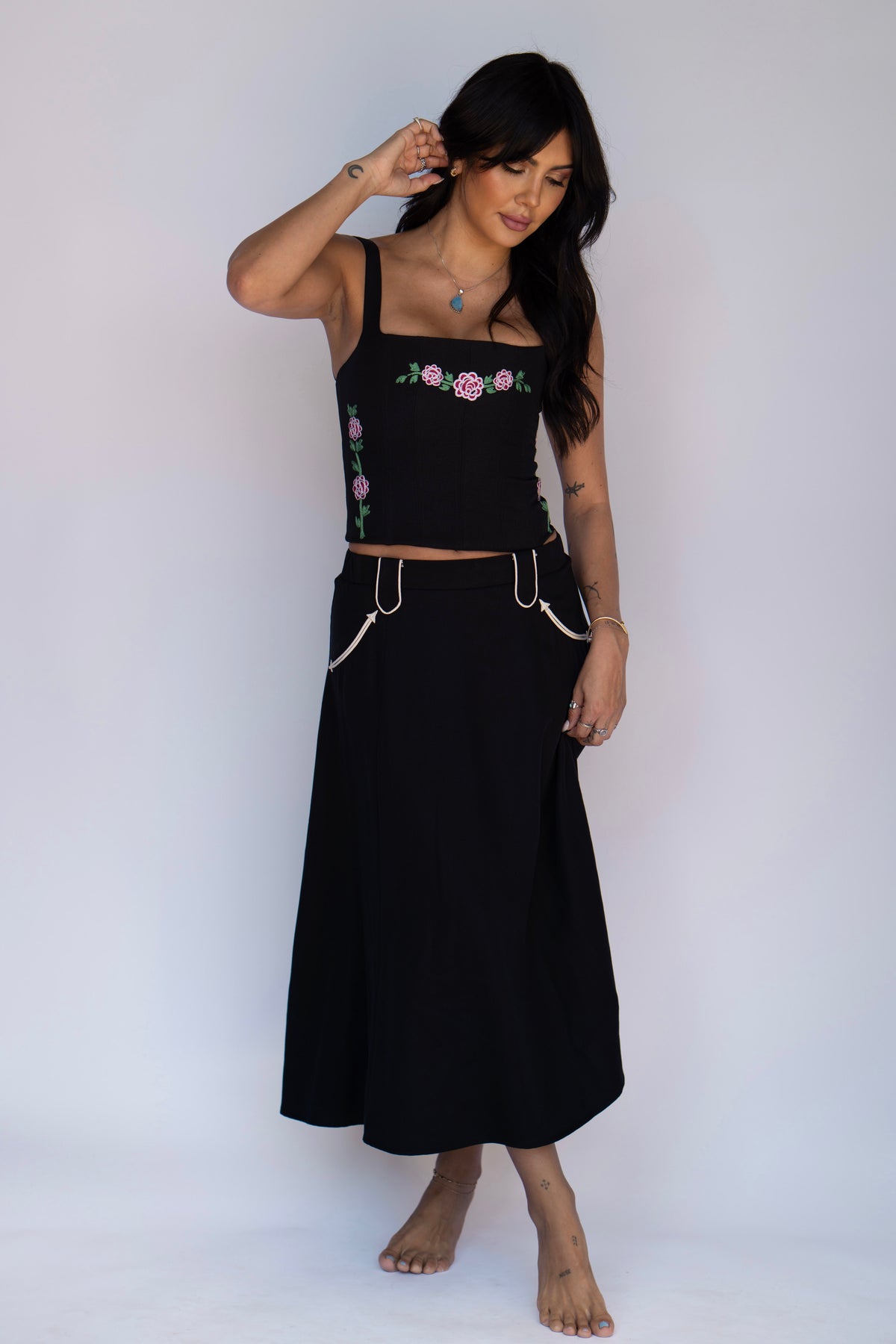 A woman modeling a black two-piece outfit consisting of a cropped tank top with pink and green floral embroidery on the chest and a matching long skirt with decorative white chain details at the waist, set against a plain white studio background.