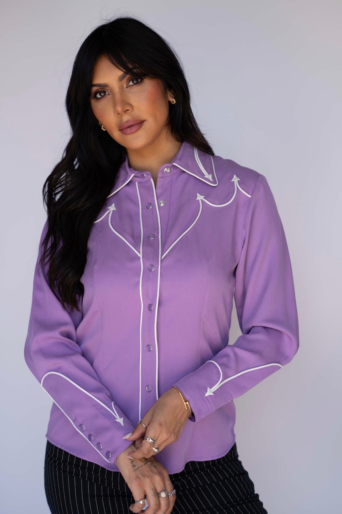 A woman modeling a lavender purple long-sleeve western-style blouse with white embroidered details and silver buttons, paired with black pinstriped pants, against a plain light gray background.