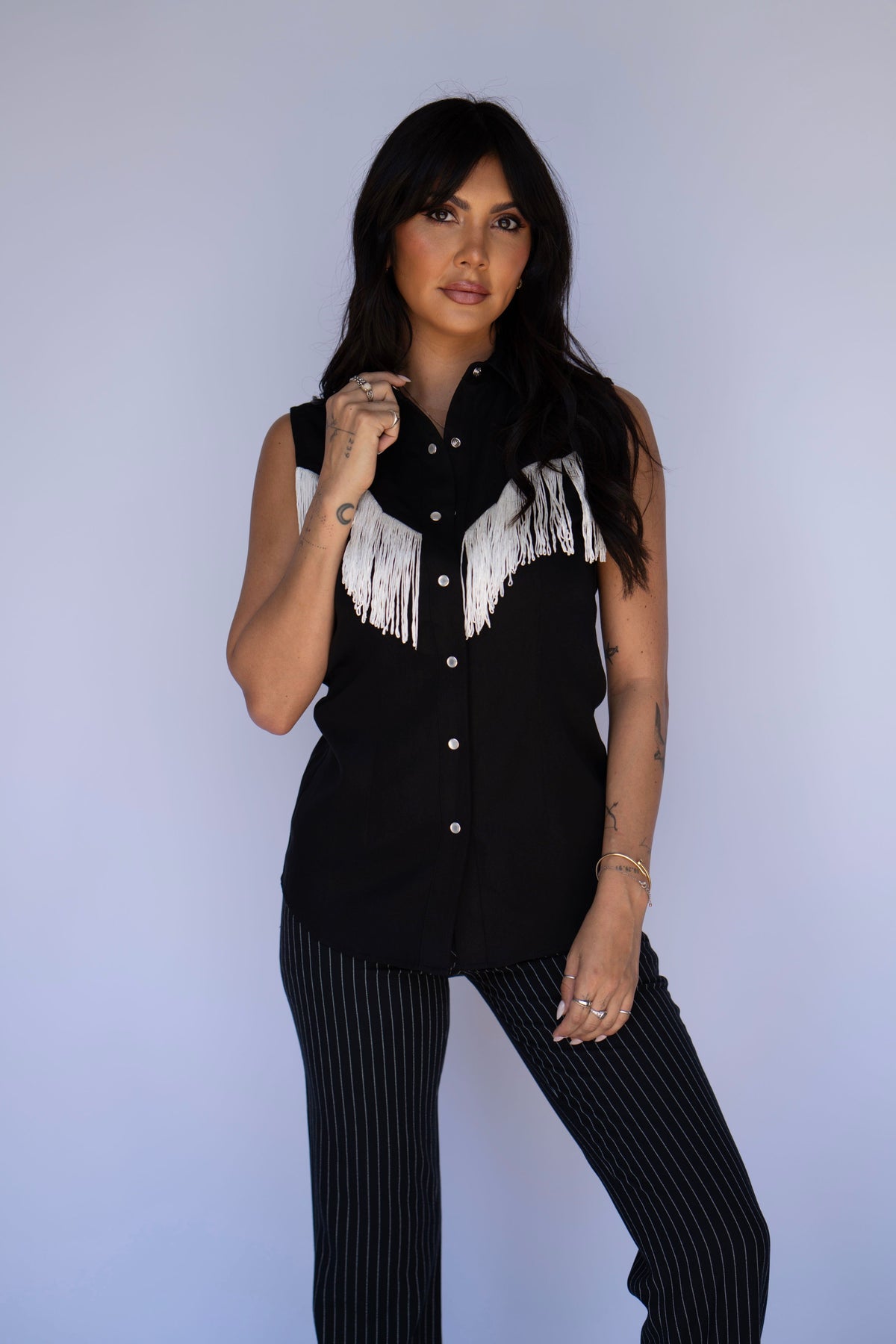 A woman modeling a sleeveless black button-up shirt with white fringe across the chest, paired with black pinstriped pants, against a light blue studio background.