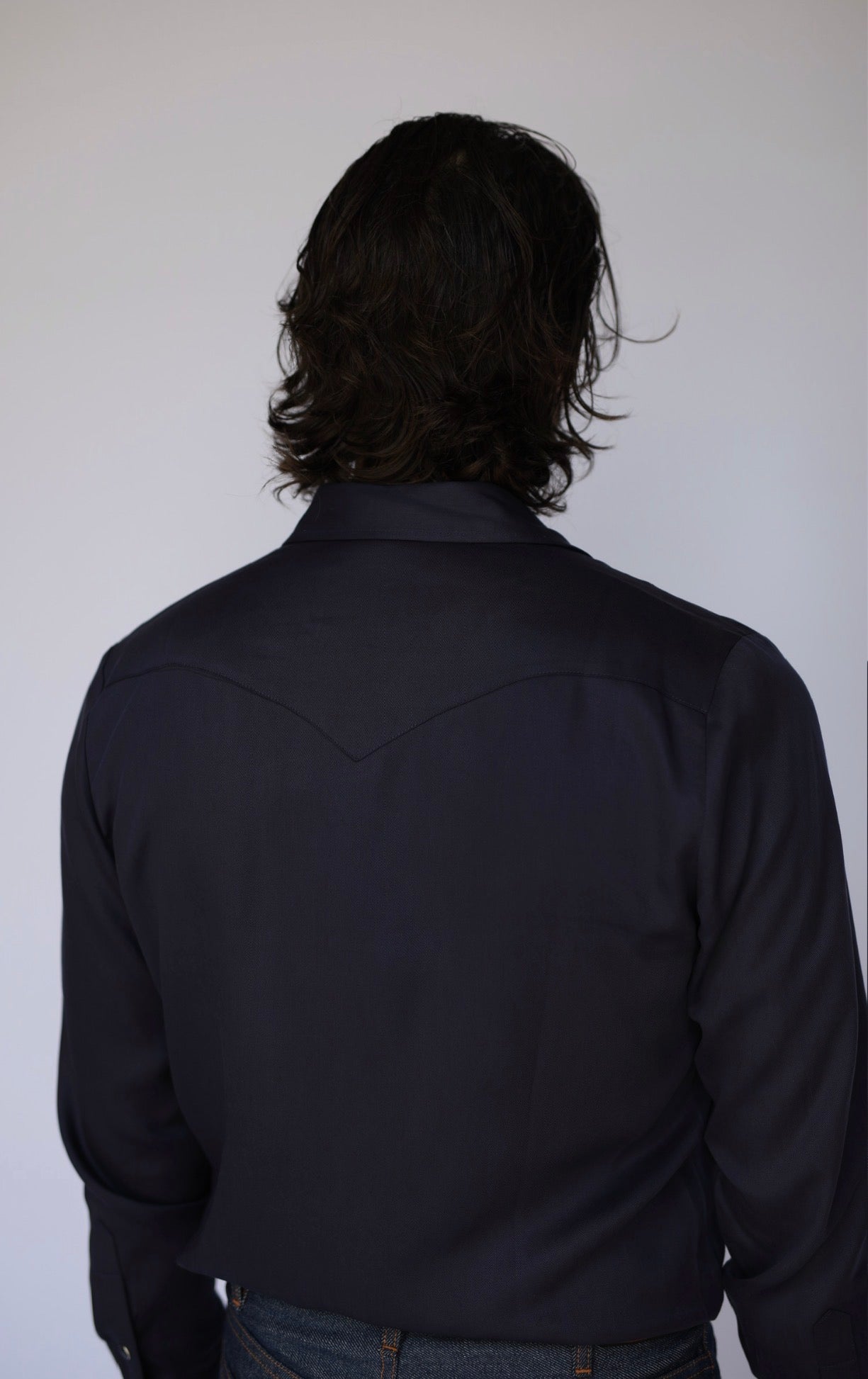 A person viewed from the back wearing a dark navy long-sleeve western-style shirt and blue jeans against a plain light gray background.