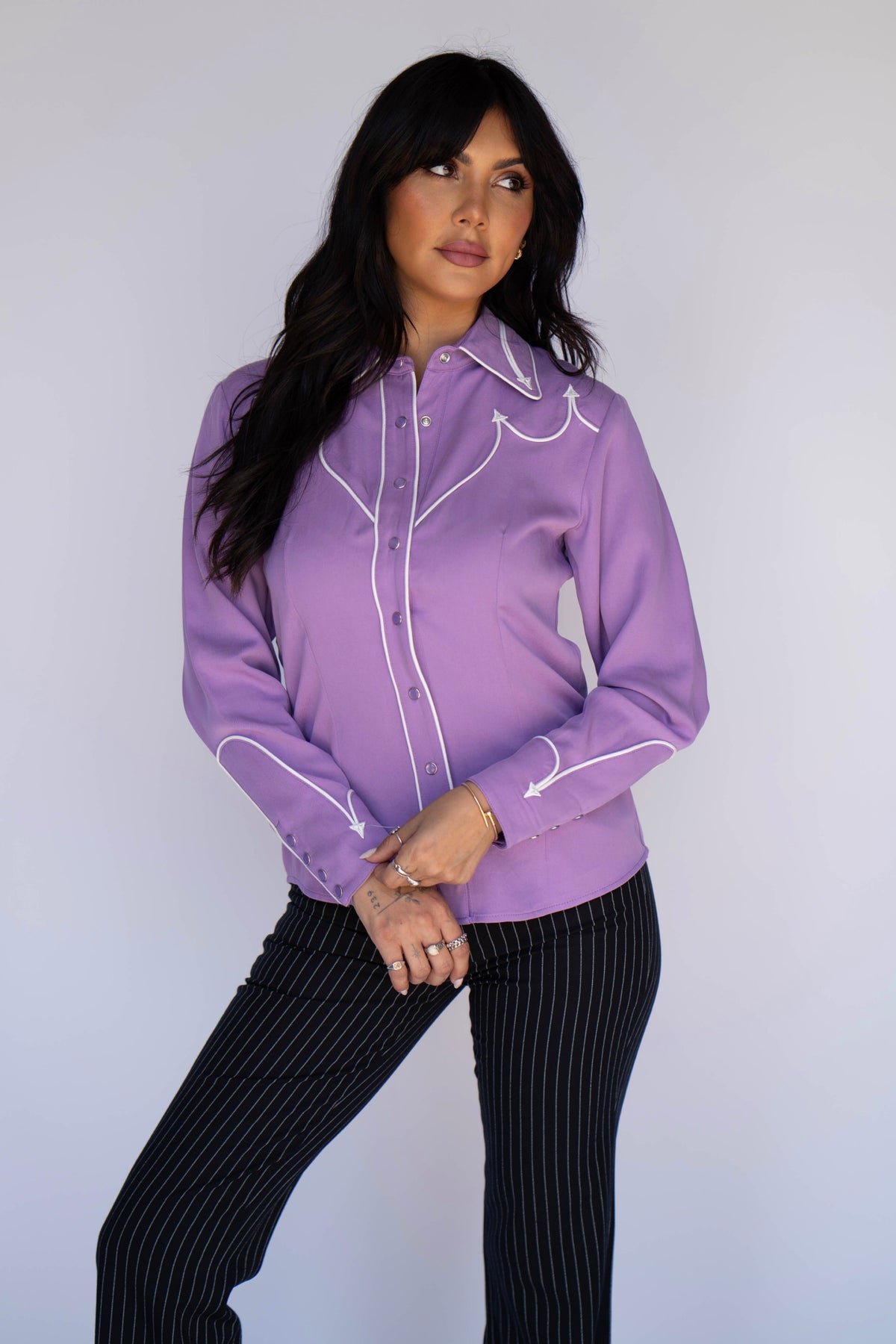 A woman modeling a lavender purple western-style long-sleeve blouse with white contrast stitching and piping, paired with black pinstriped pants, against a plain light gray studio background.