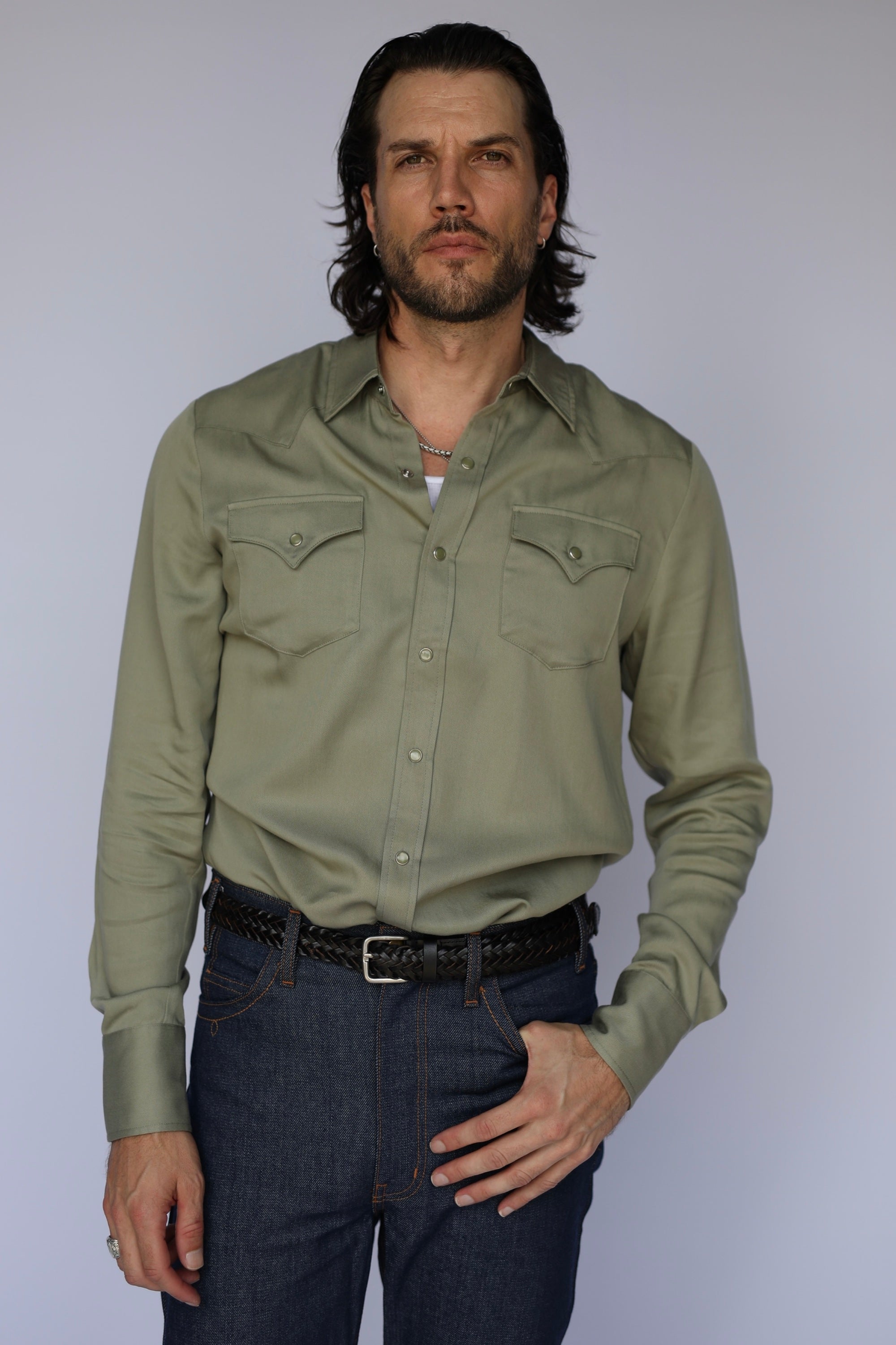 A man modeling a sage green western-style long-sleeve button-down shirt with two chest pockets, paired with dark blue jeans and a black belt, against a light gray studio background.