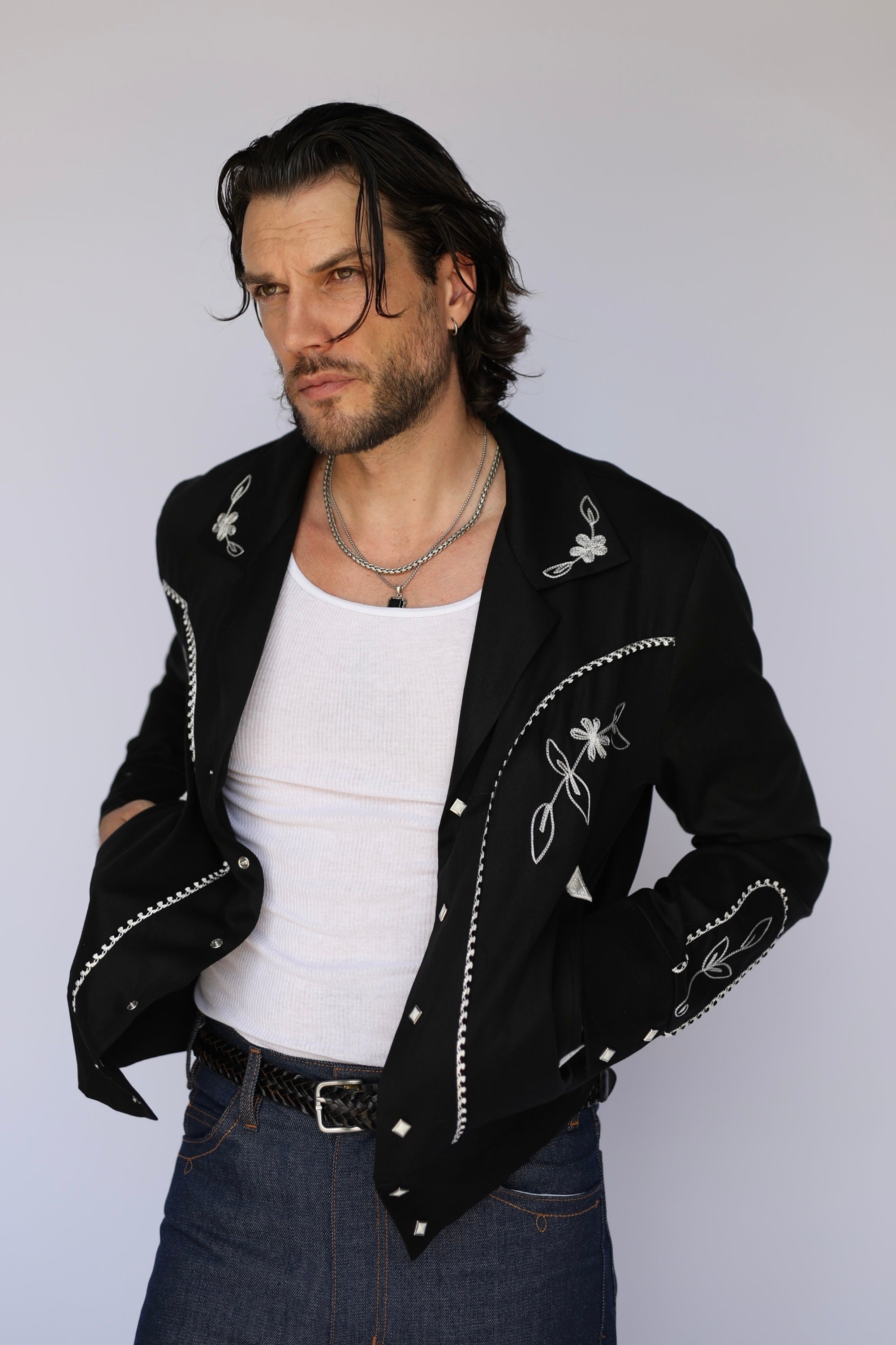 A man wearing a black western-style jacket with white embroidered details and silver studs, layered over a white tank top and blue jeans, photographed against a light gray studio background.