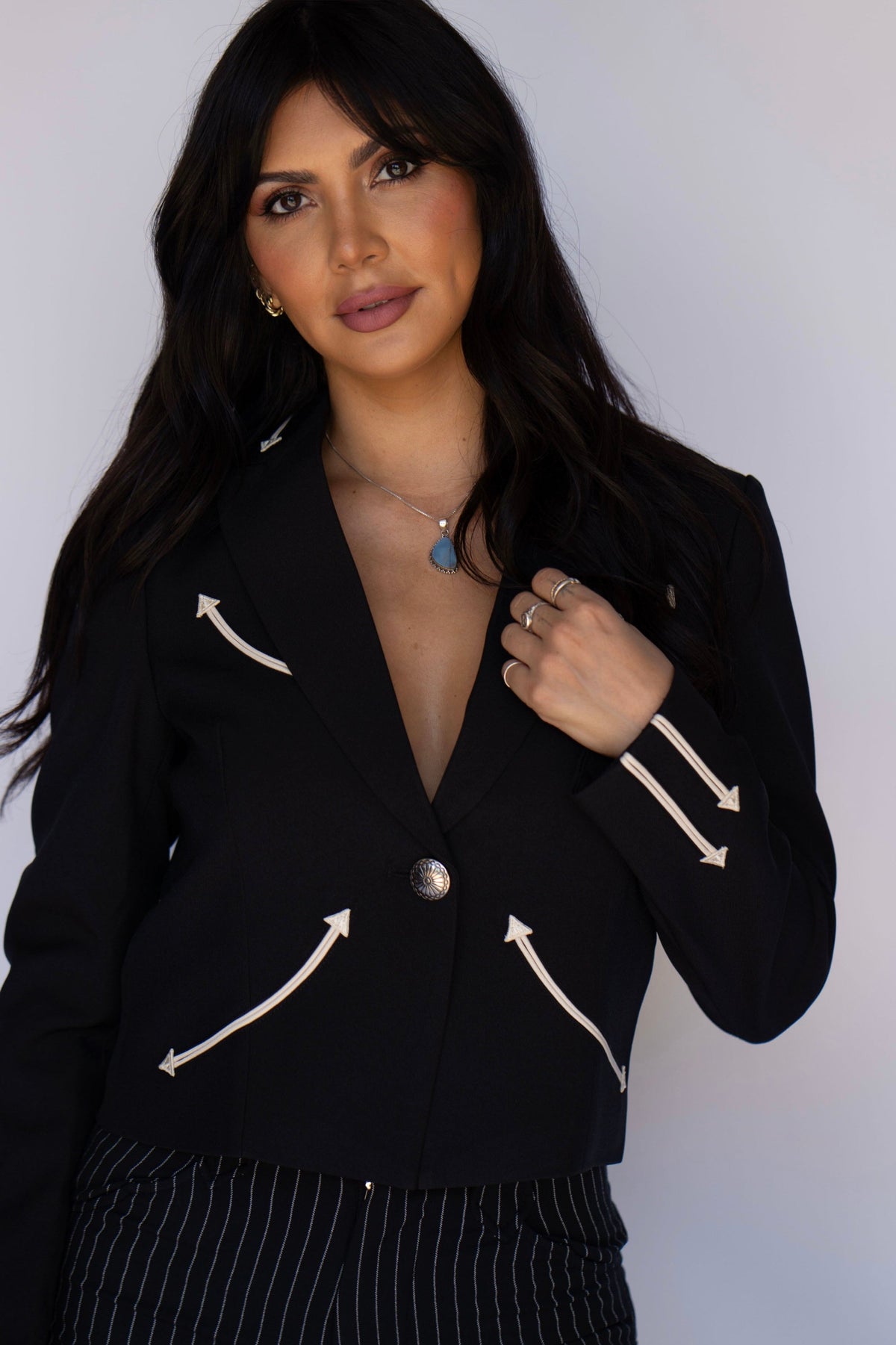 A woman wearing a black cropped blazer with white arrow details on the chest and sleeves, paired with black pinstriped trousers, against a plain light gray background.