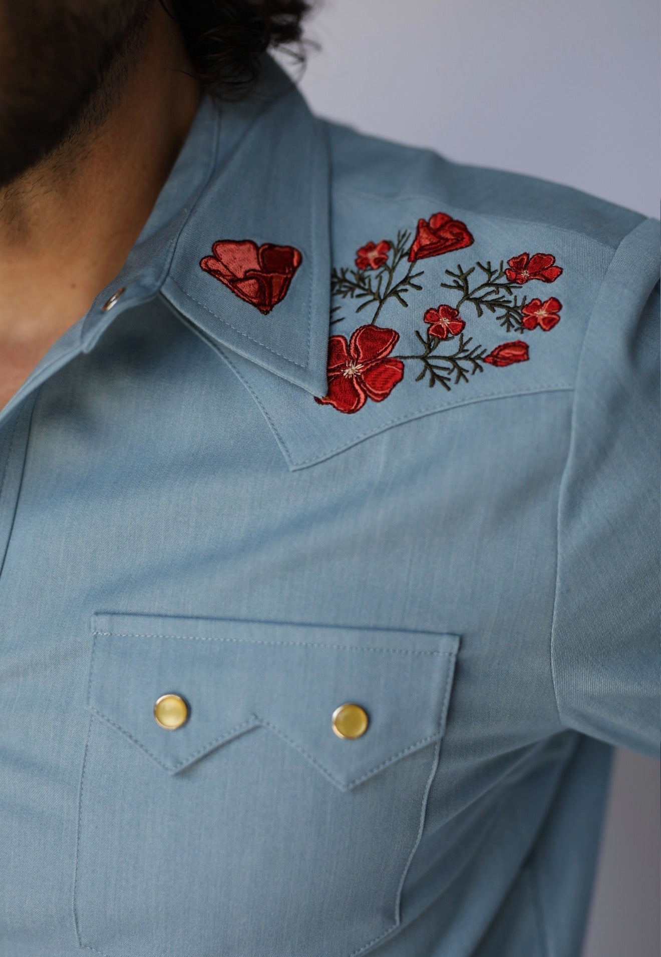 Close-up of a light blue western-style shirt with red floral embroidery on the chest and gold snap buttons on a chest pocket.