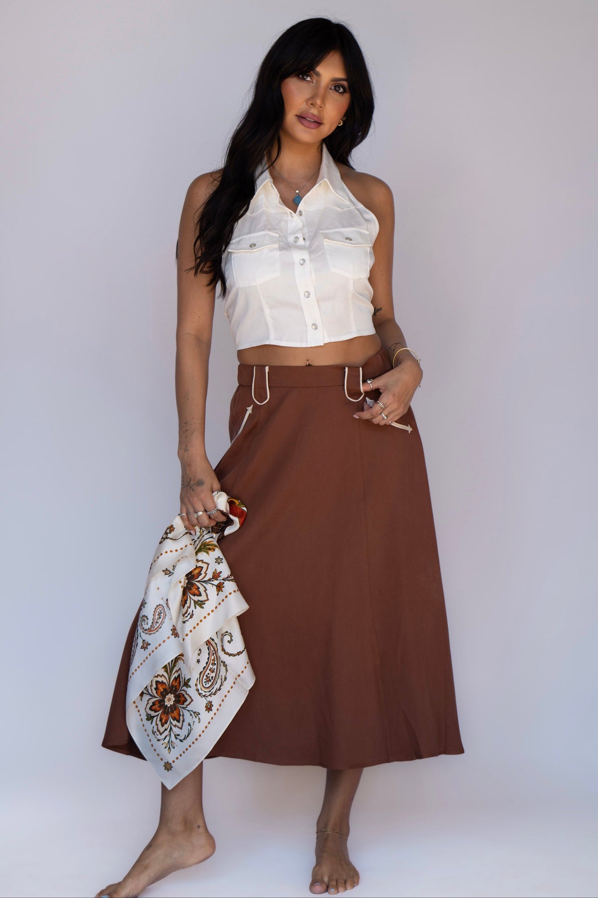 A woman stands barefoot against a light gray studio background, wearing a white sleeveless cropped button-front top and a brown midi skirt with a drawstring waist. She holds a white paisley bandana in one hand.