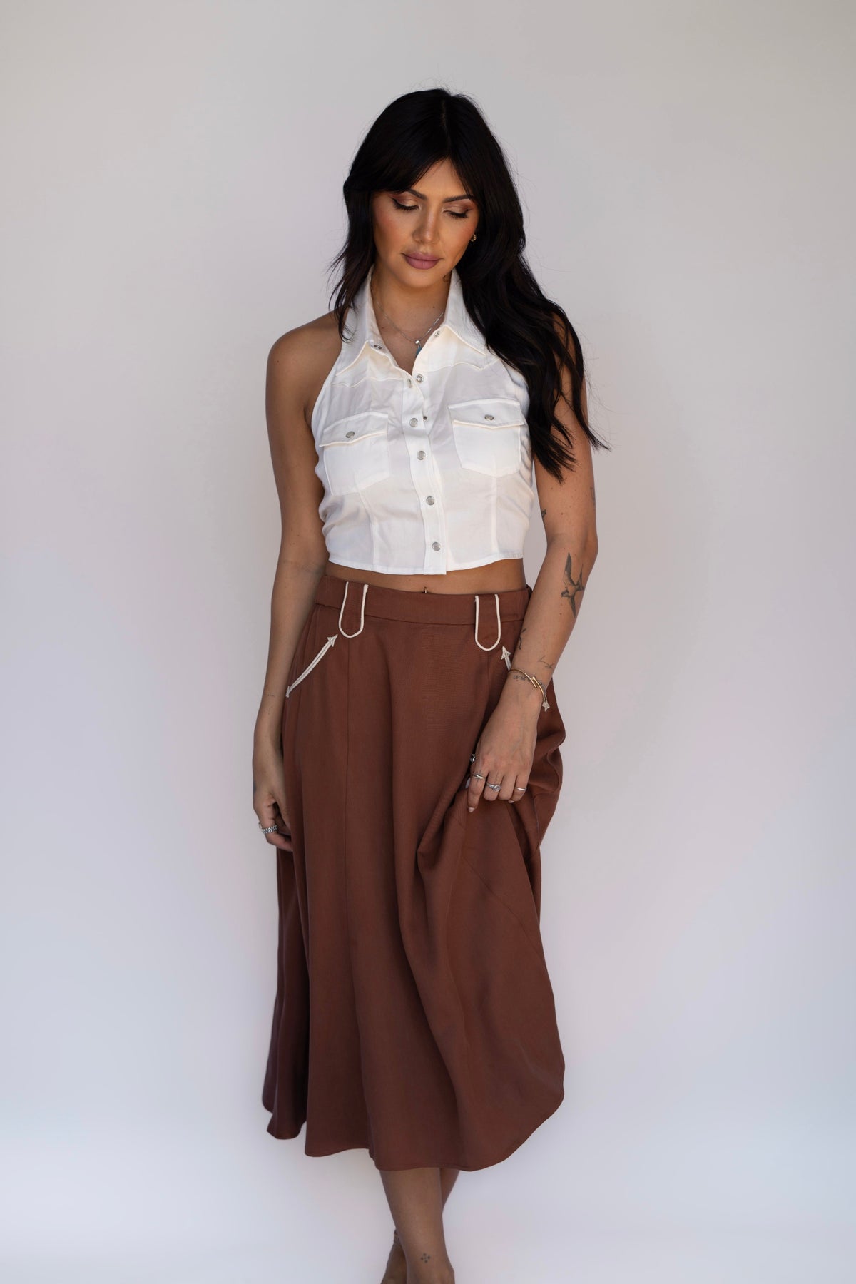 A woman modeling a white sleeveless cropped button-up shirt and a brown midi skirt with white drawstrings, standing against a plain light gray studio background.