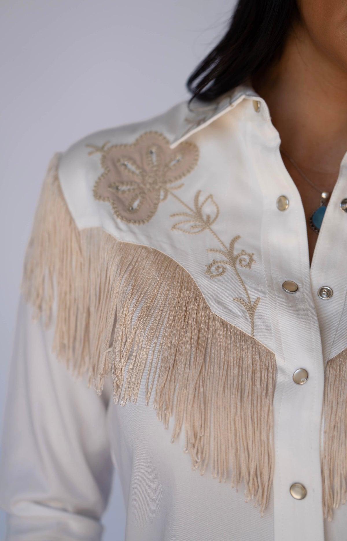 Close-up of a white western-style blouse with beige fringe, embroidered floral detailing on the yoke, and gold-tone buttons, worn by a model.