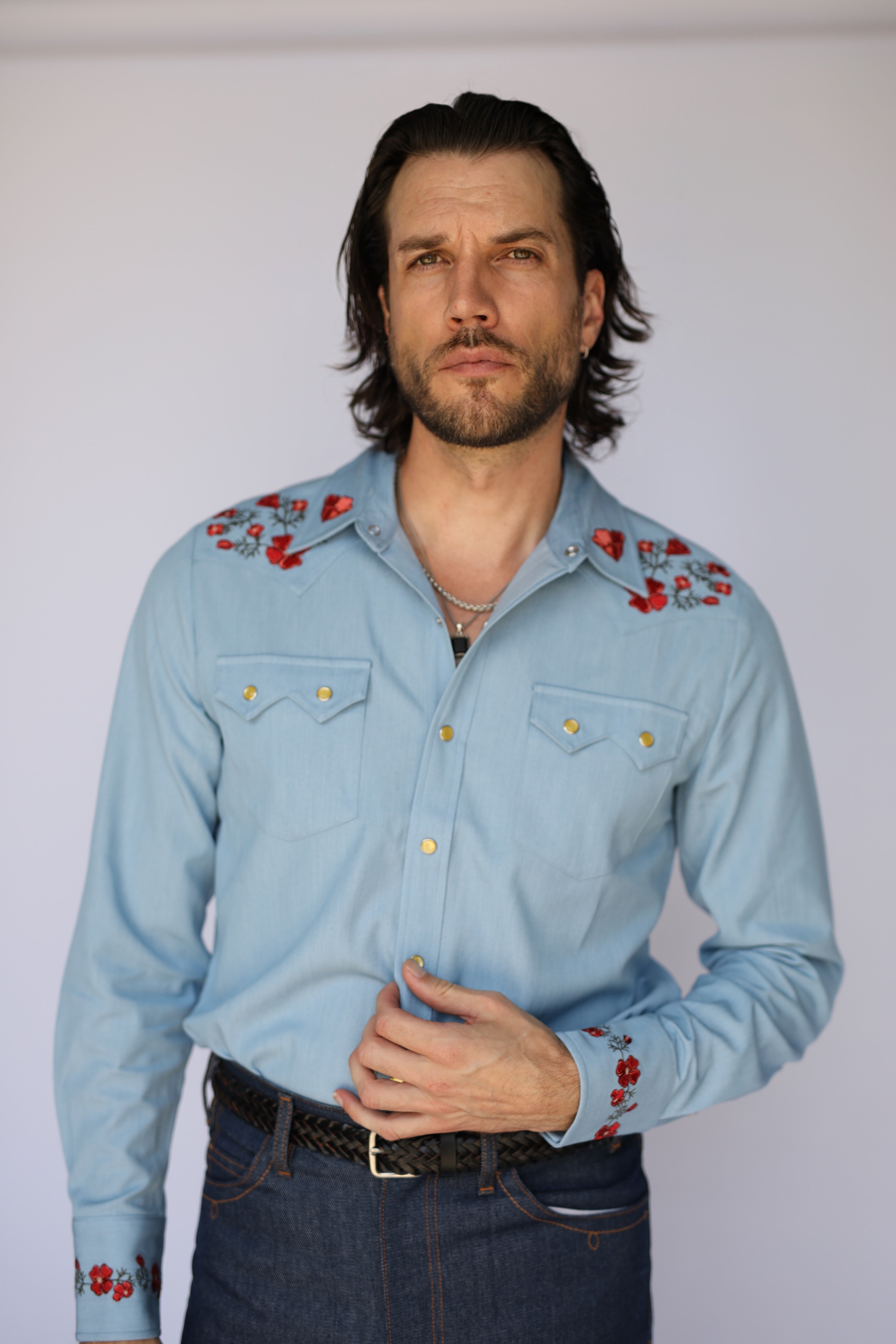 A man wearing a light blue western-style long-sleeve shirt with red and white floral embroidery on the shoulders and cuffs, paired with dark blue jeans and a black belt, against a plain light background.