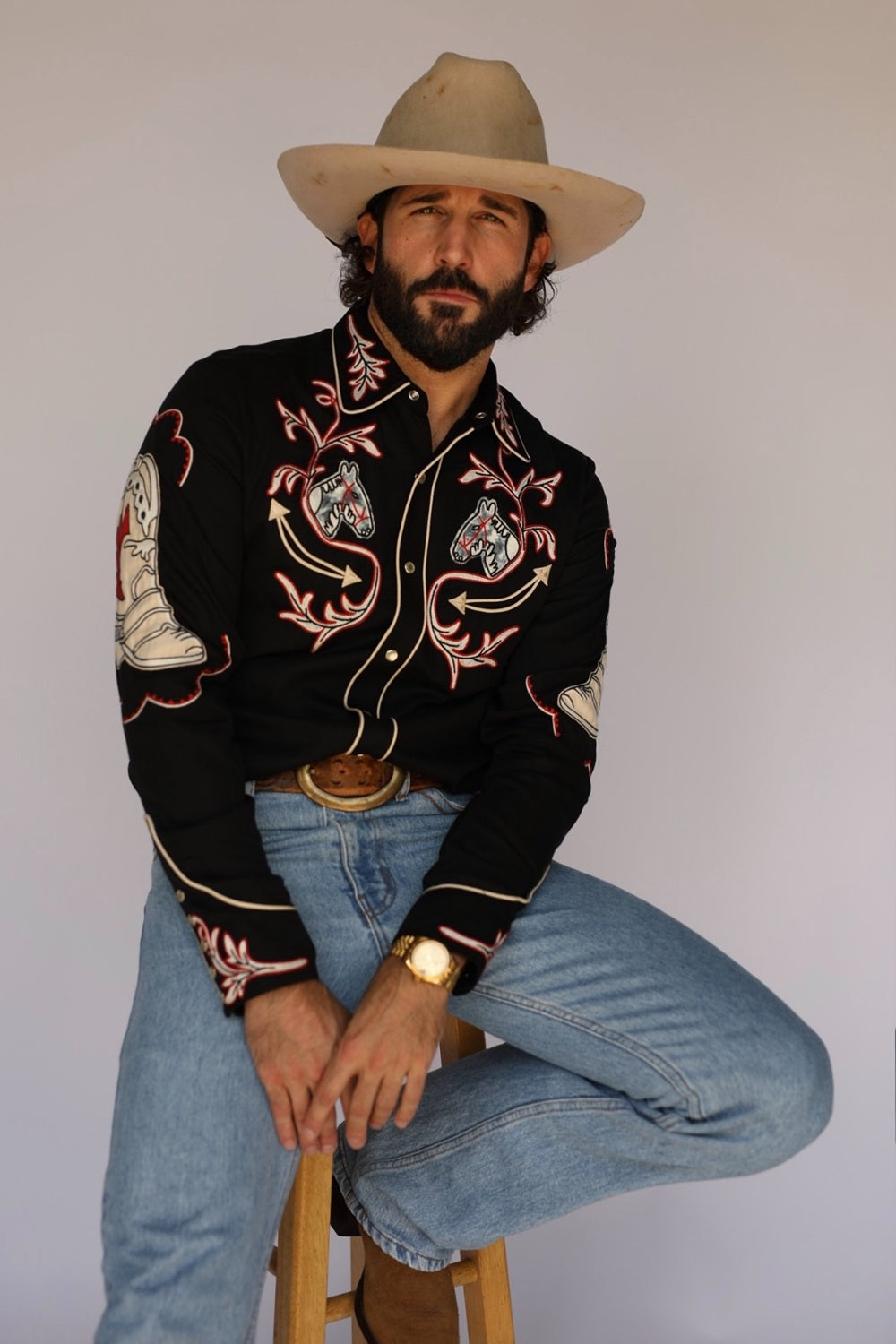 A man wearing a black western-style embroidered shirt, blue jeans, a tan cowboy hat, and a brown belt, sitting on a wooden stool against a plain light gray background.