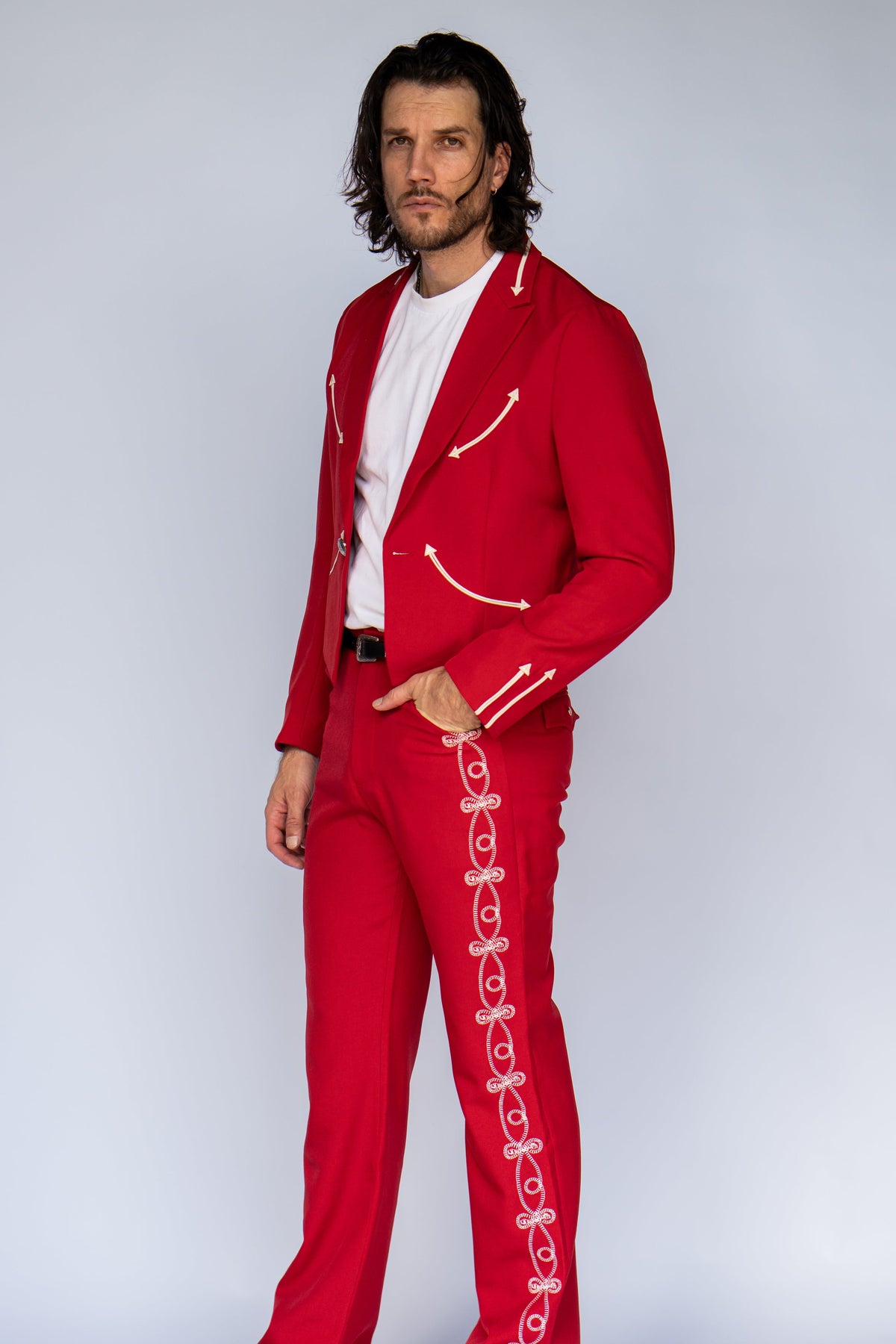 A man modeling a bright red two-piece tracksuit with white decorative piping and side details, worn over a white t-shirt and white slip-on shoes, against a light gray studio background.