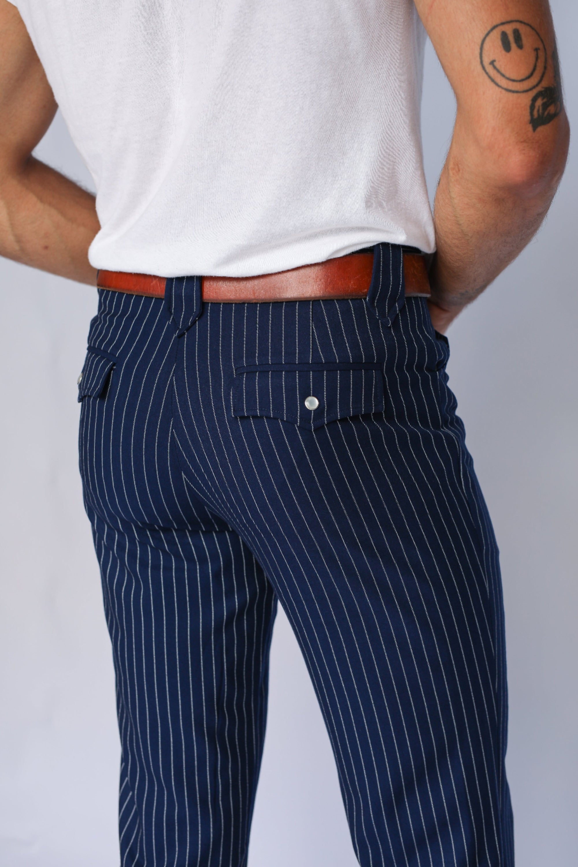 A man wearing navy blue pinstripe trousers with a brown leather belt and a white tank top, shown from the back against a light gray background.