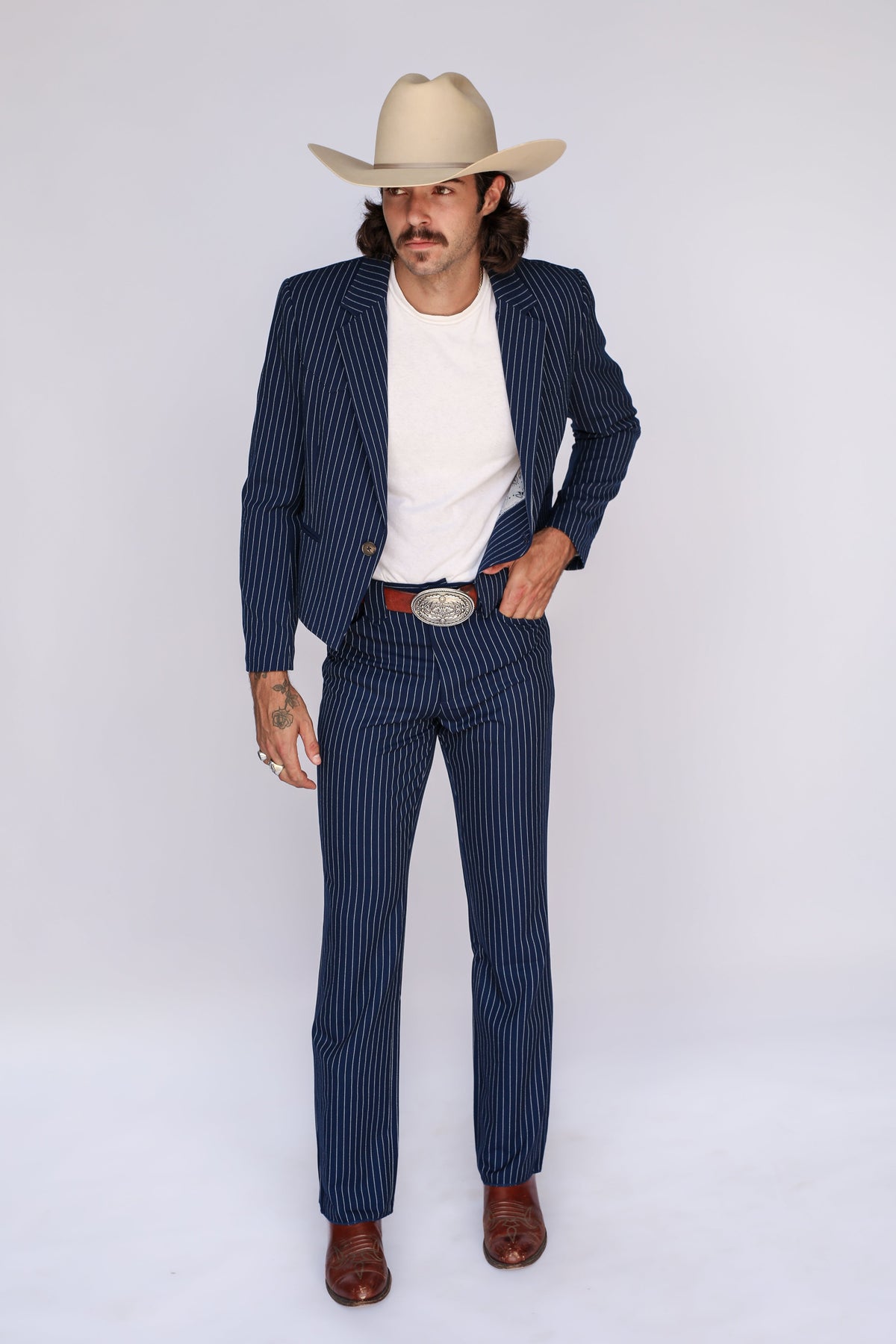 A man modeling a navy blue pinstripe suit with a white t-shirt, brown leather boots, a red belt, and a beige cowboy hat, standing against a white studio background.