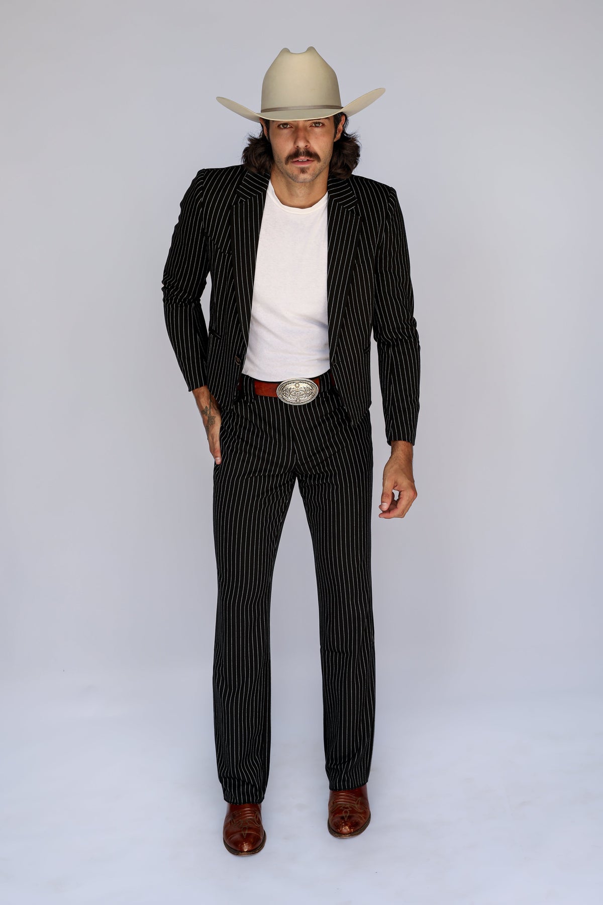 A man modeling a black pinstripe suit with a white t-shirt, brown leather belt, silver buckle, and a beige cowboy hat, standing against a light gray studio background.