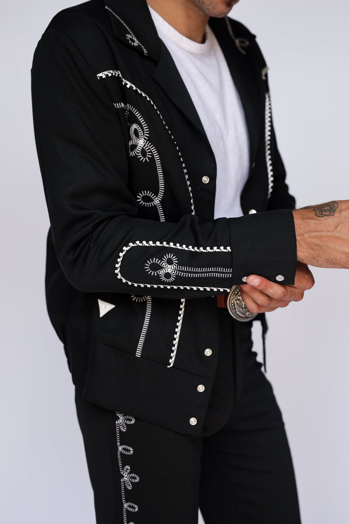 A close-up of a person wearing a black western-style blazer with white embroidered detailing, paired with black pants and a white t-shirt, against a light gray background.
