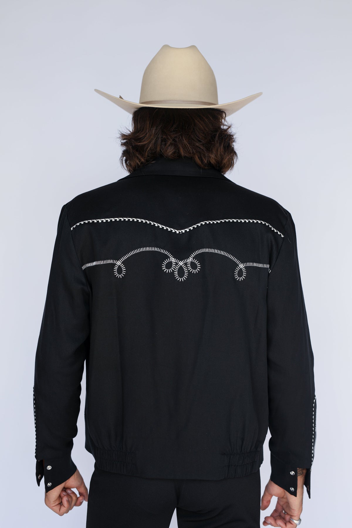 A man viewed from behind wearing a black western-style jacket with silver embroidered scrollwork across the back and a light beige cowboy hat, against a light gray studio background.