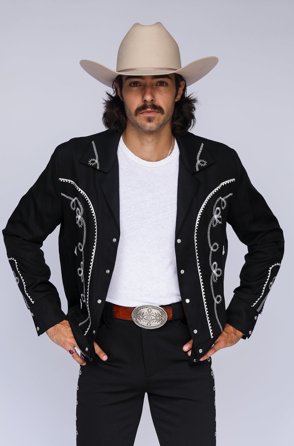 A man modeling a black western-style jacket and matching pants with silver embroidery, a white t-shirt, a brown belt with a large silver buckle, brown leather shoes, and a beige cowboy hat, standing against a light gray studio background.